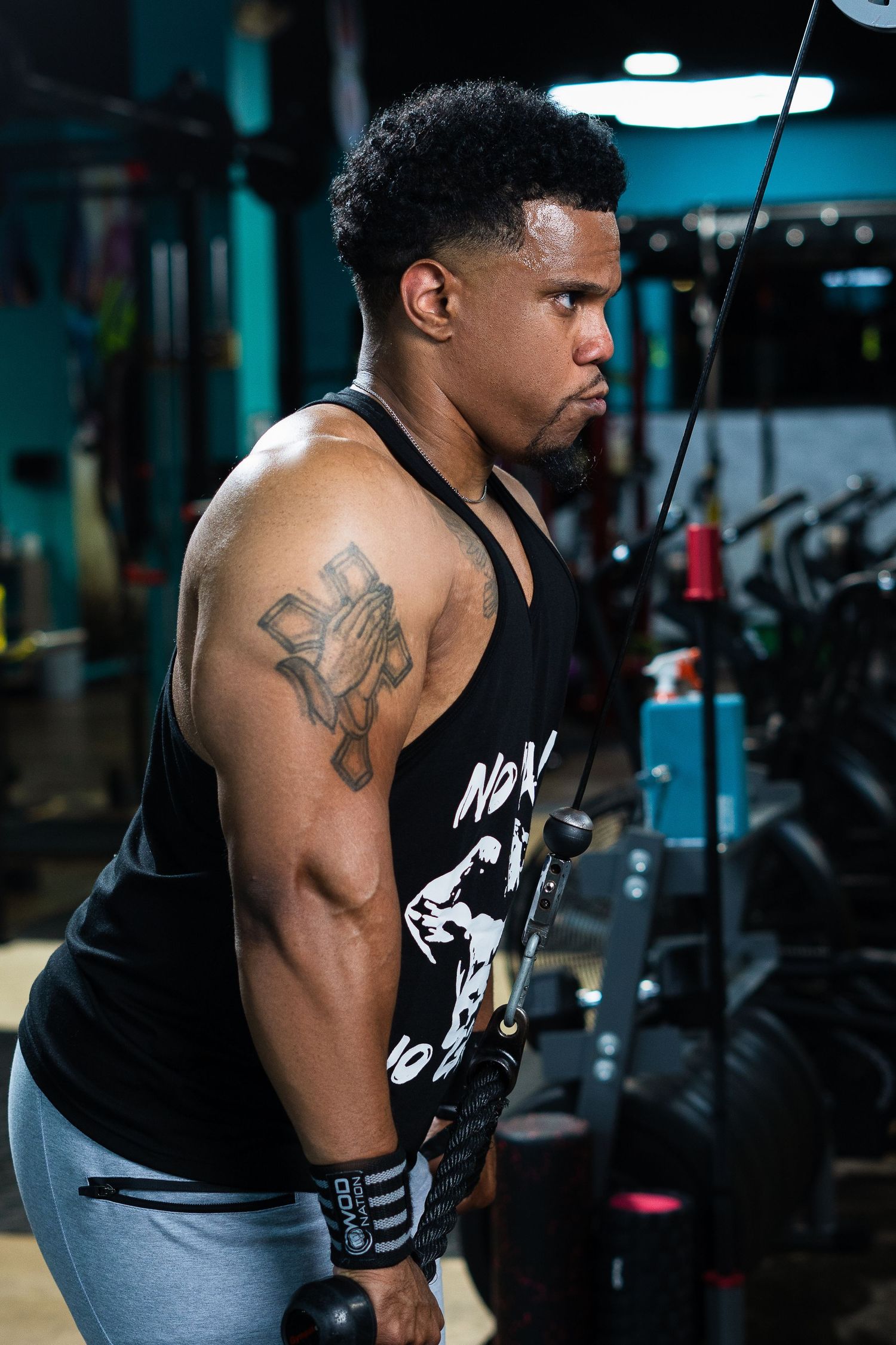 Person doing cable machine exercises wearing black tank top and gray pants in gym.