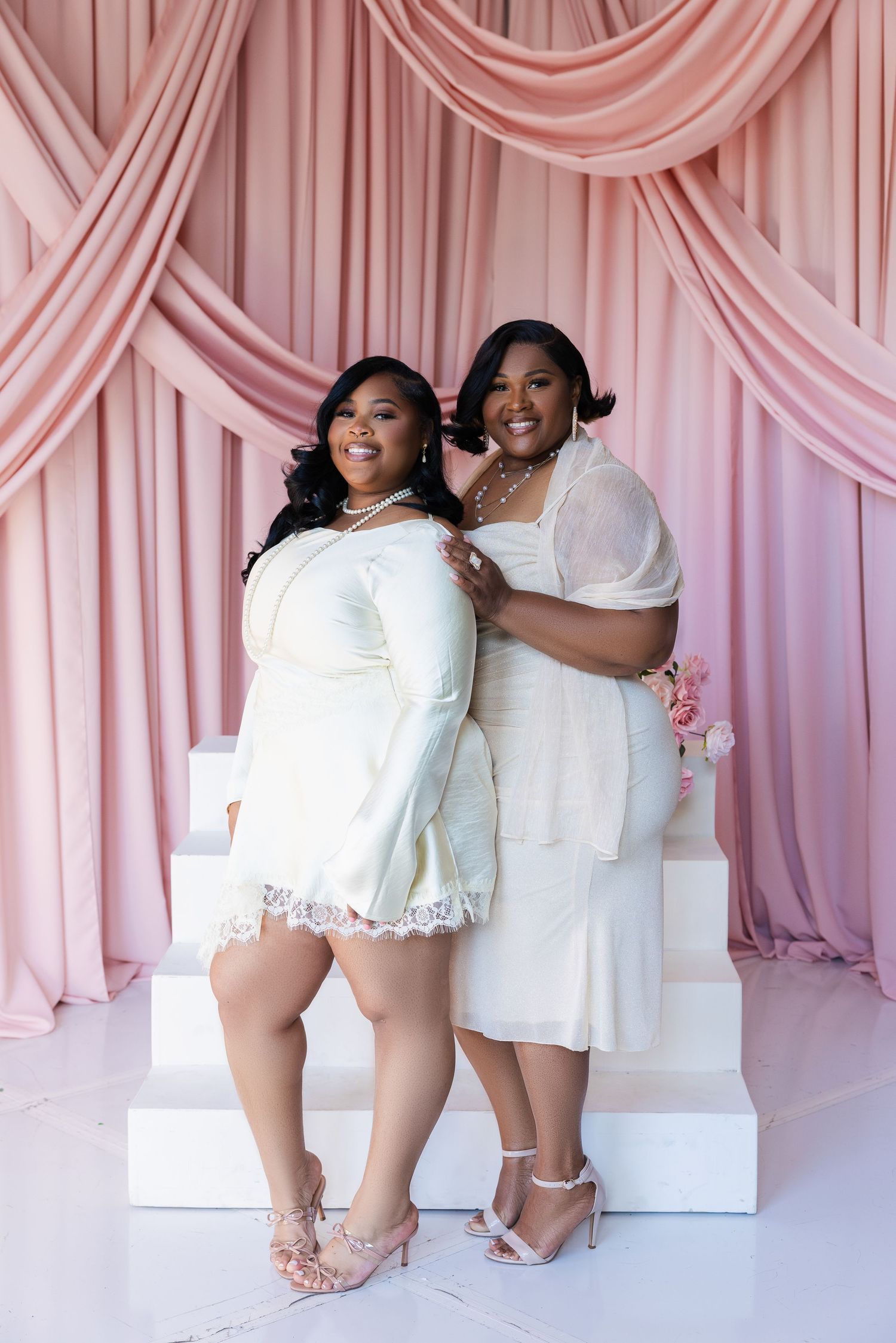 Two women in white dresses pose elegantly against pink draped backdrop with floral decor.