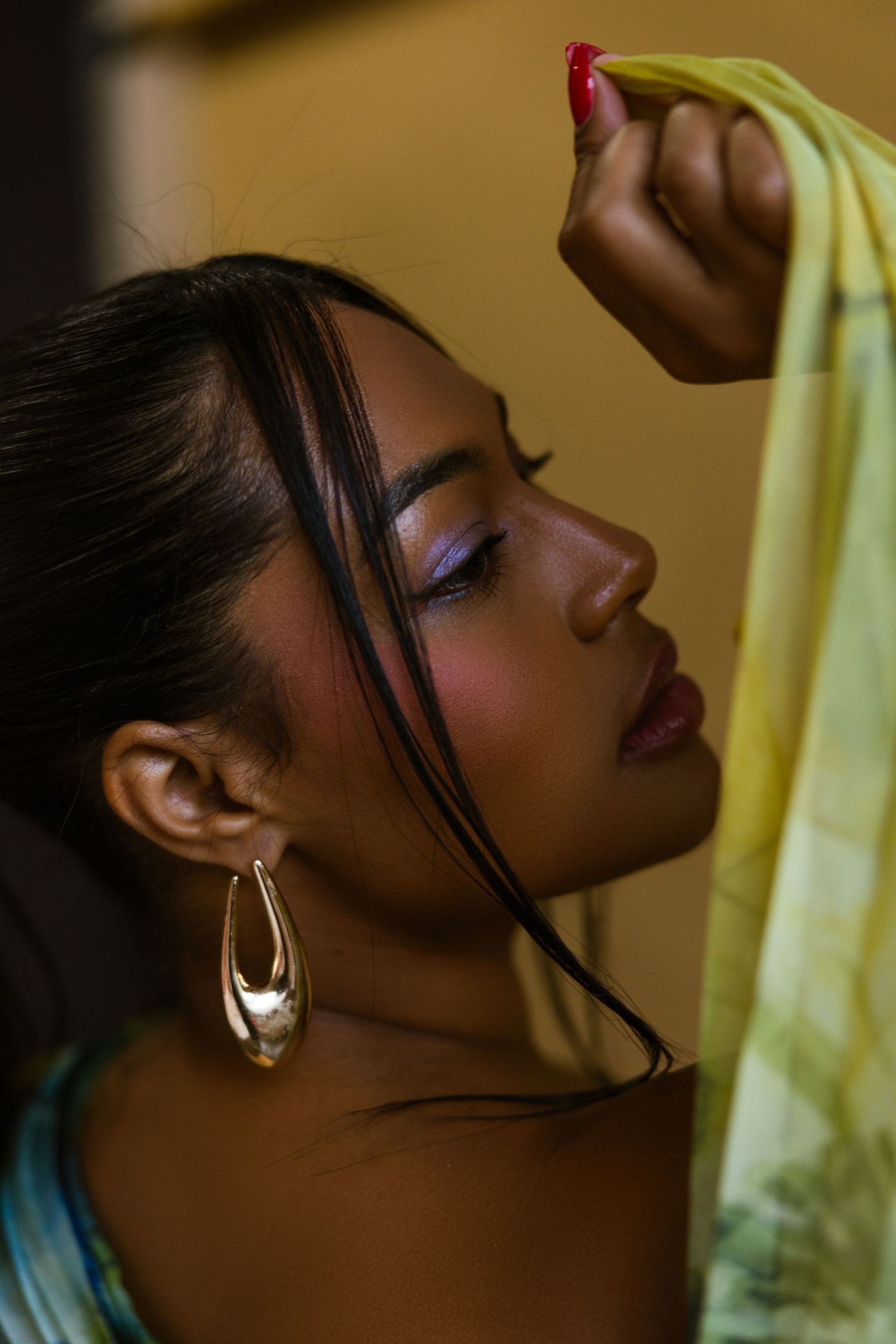 A woman with purple eyeshadow and gold hoop earrings holds a yellow fabric, eyes closed, in a artistic portrait.