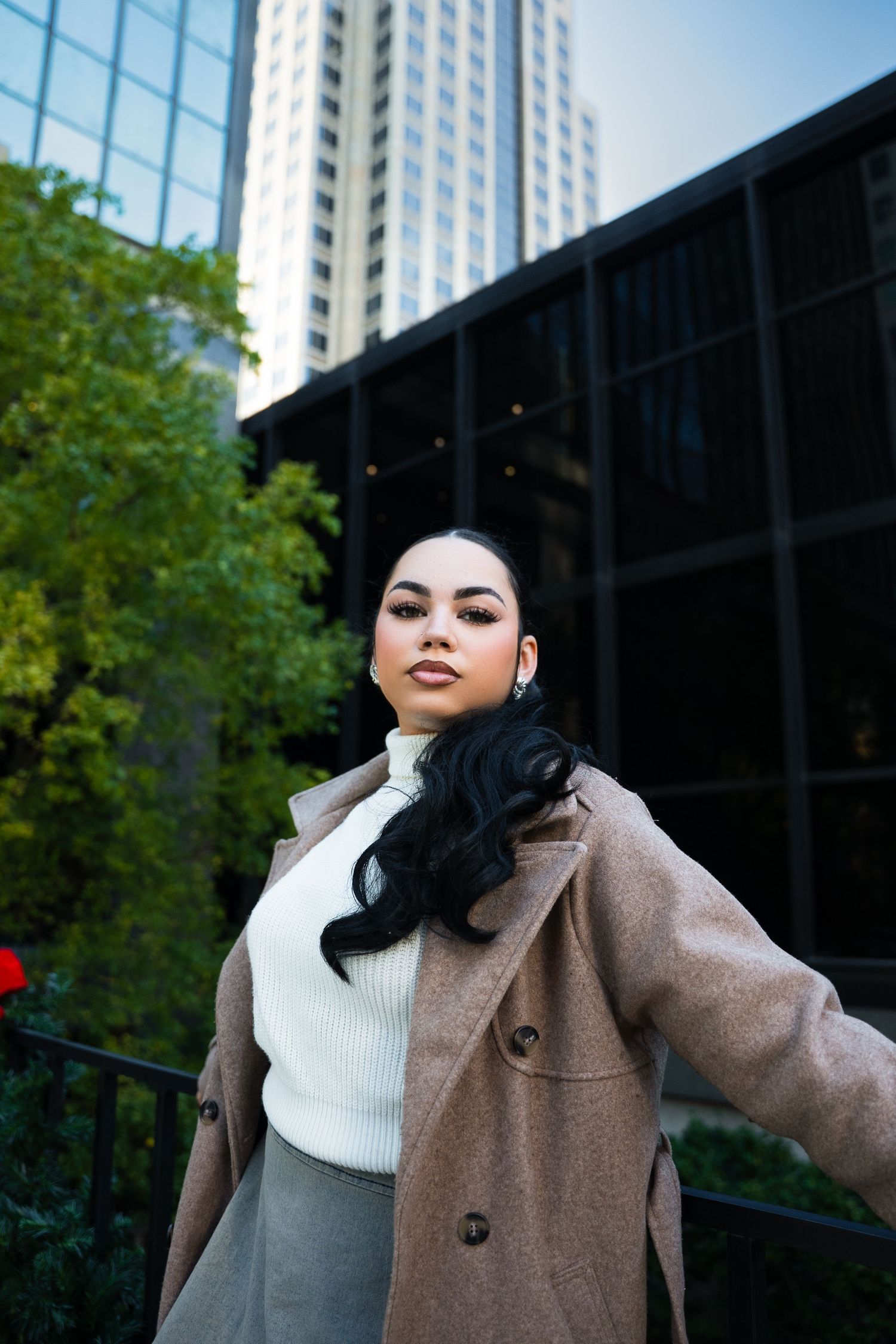 Fashion portrait of someone in a beige coat and white sweater posing against an urban backdrop with tall buildings.