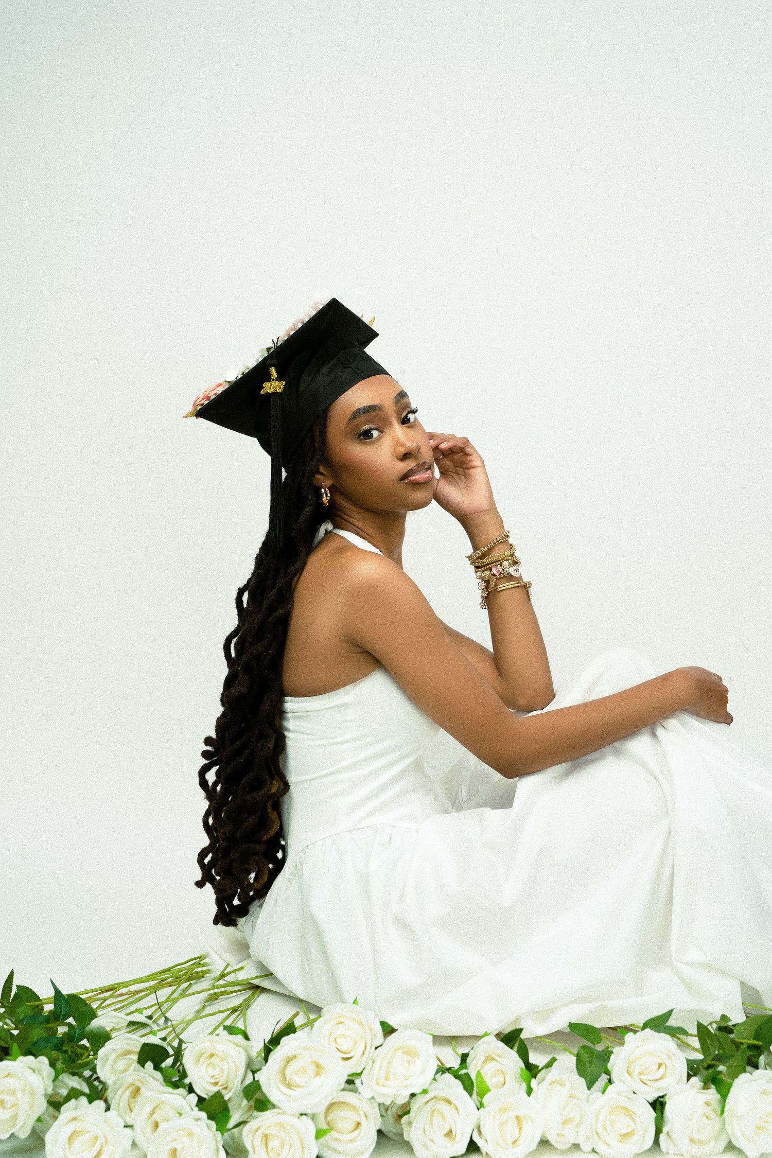 Graduate in white dress sits elegantly among white roses wearing a graduation cap and long curly hair.
