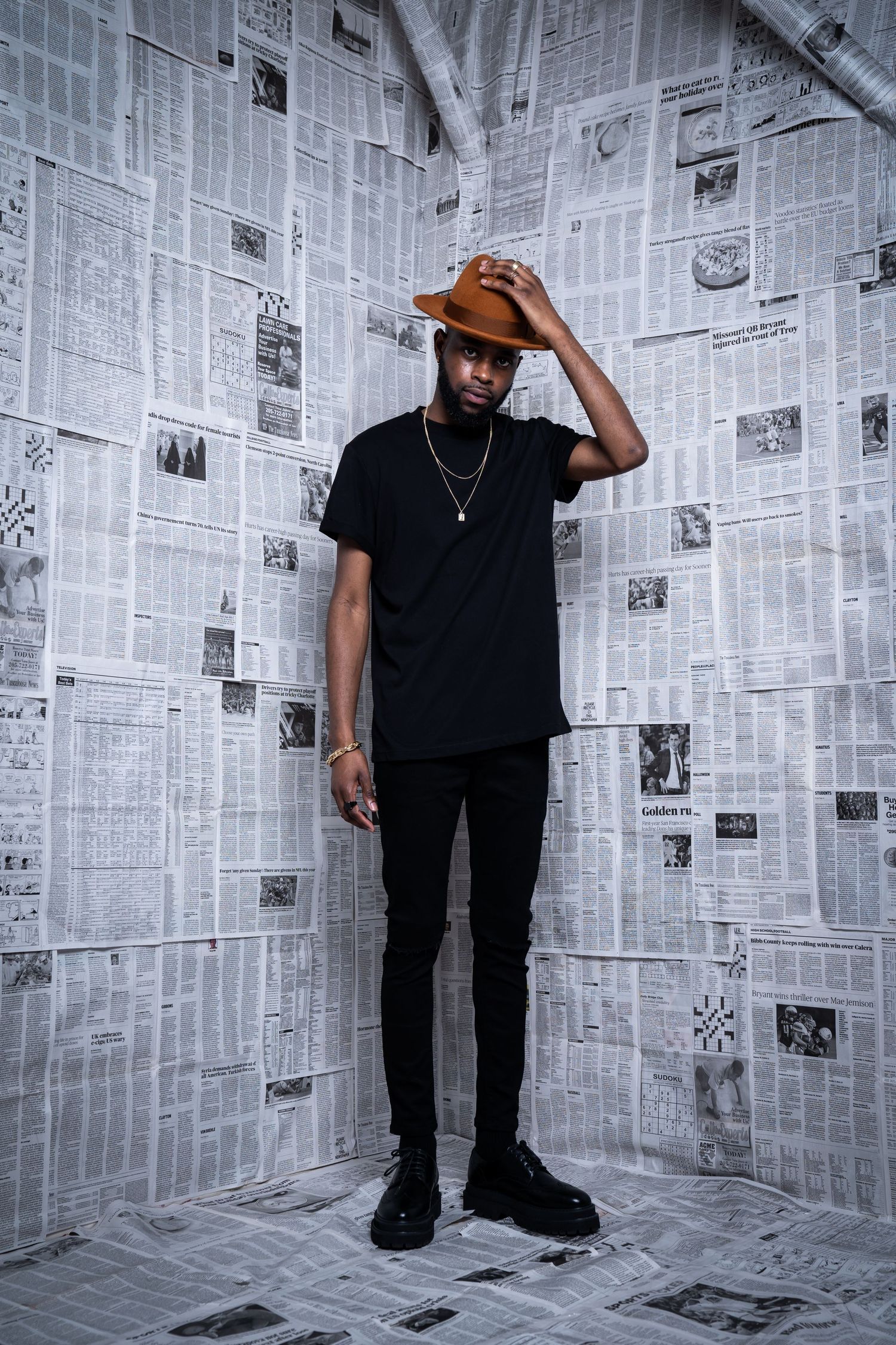 Person in black outfit and brown hat poses against wall covered in newspaper clippings.