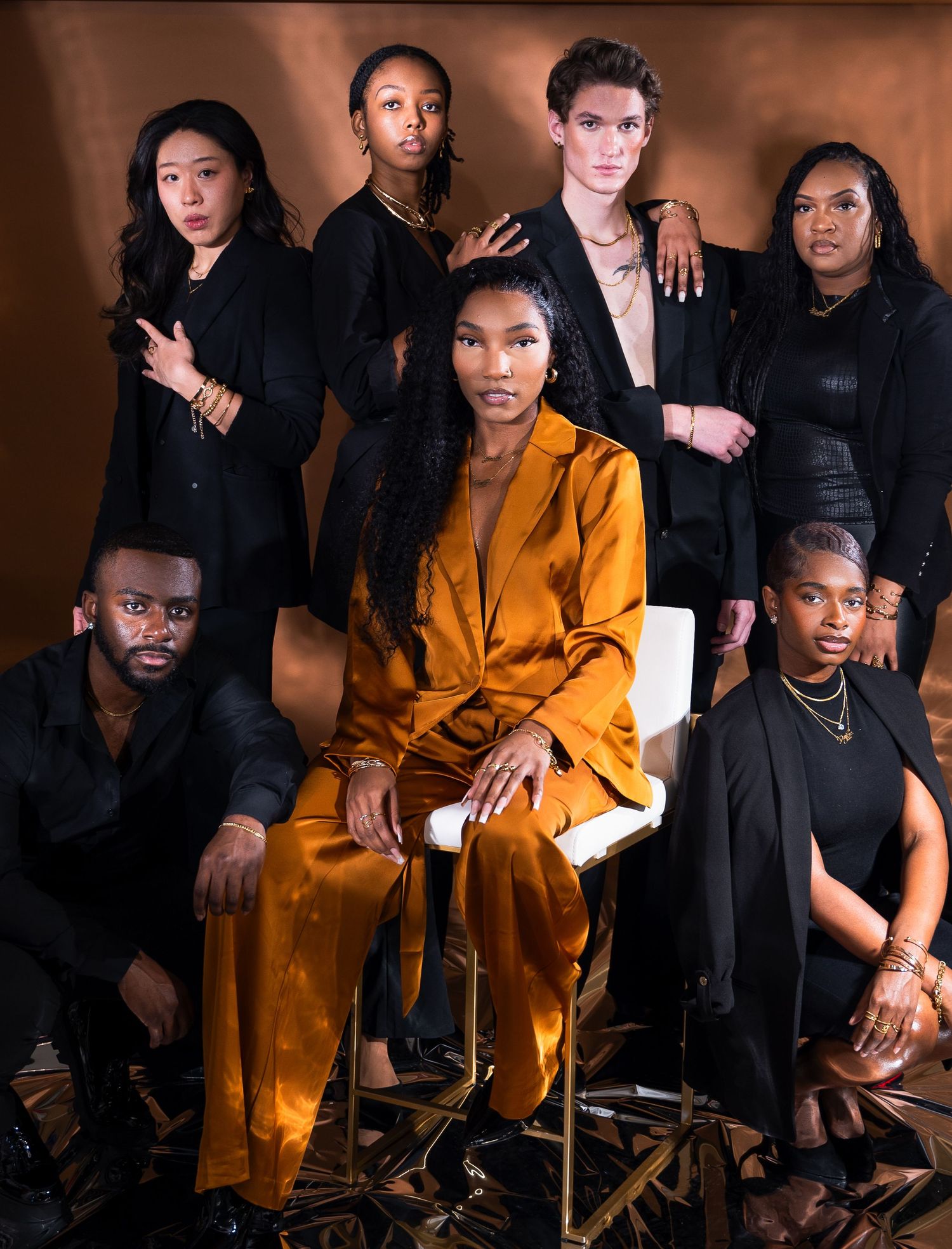 Group fashion portrait with person in gold suit surrounded by others in black attire.
