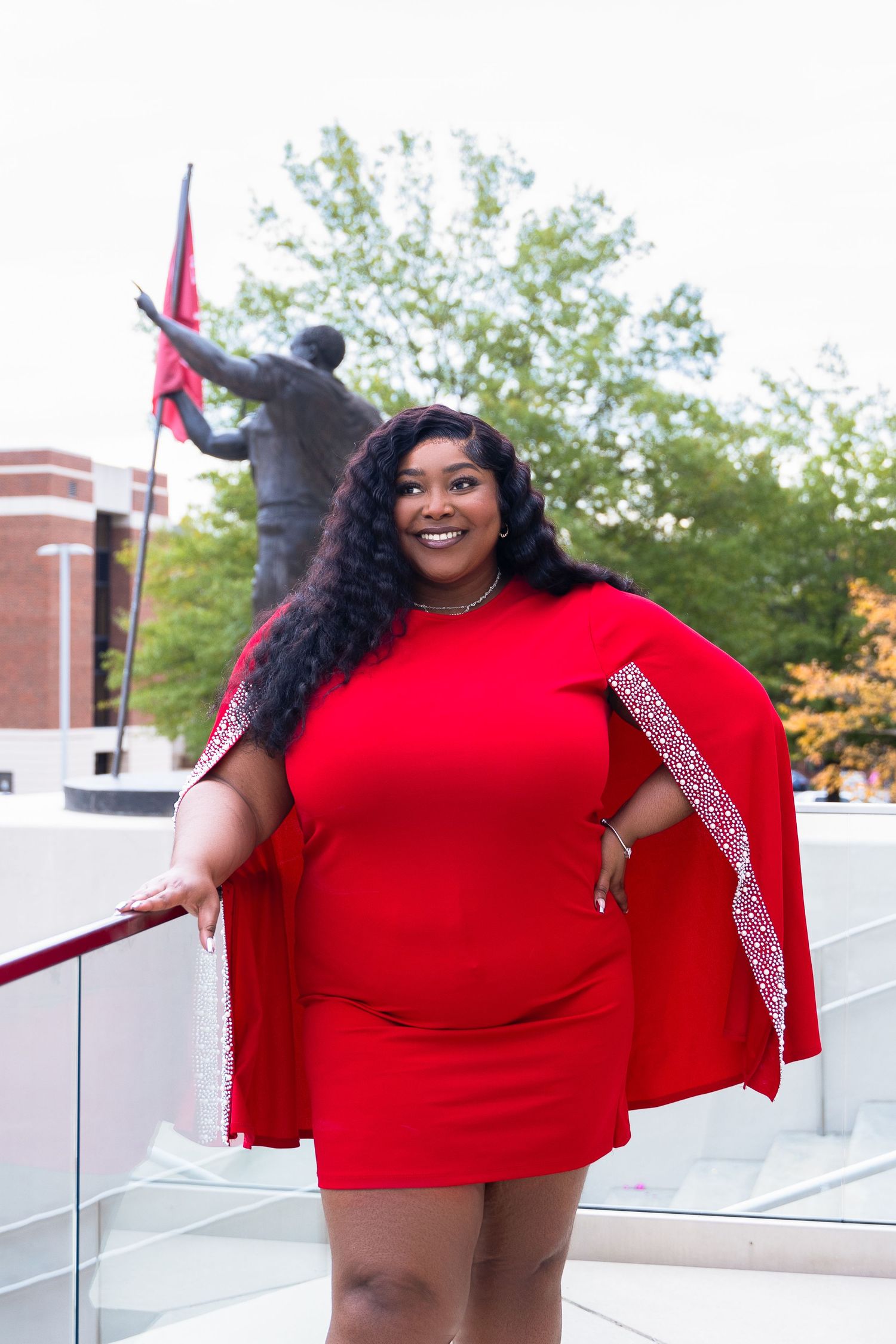 Cheerful graduate in red dress with cape standing outdoors near campus architecture.