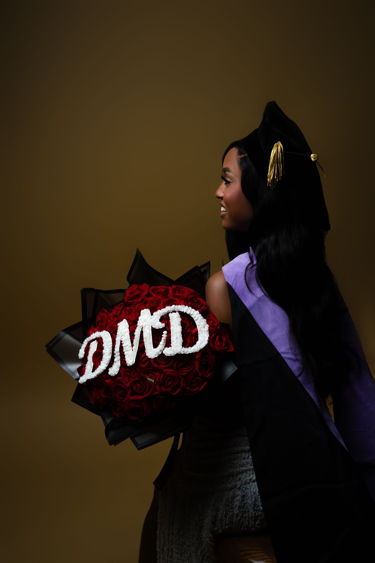 Graduate in cap and gown holds red rose bouquet spelling DMD against dark background.