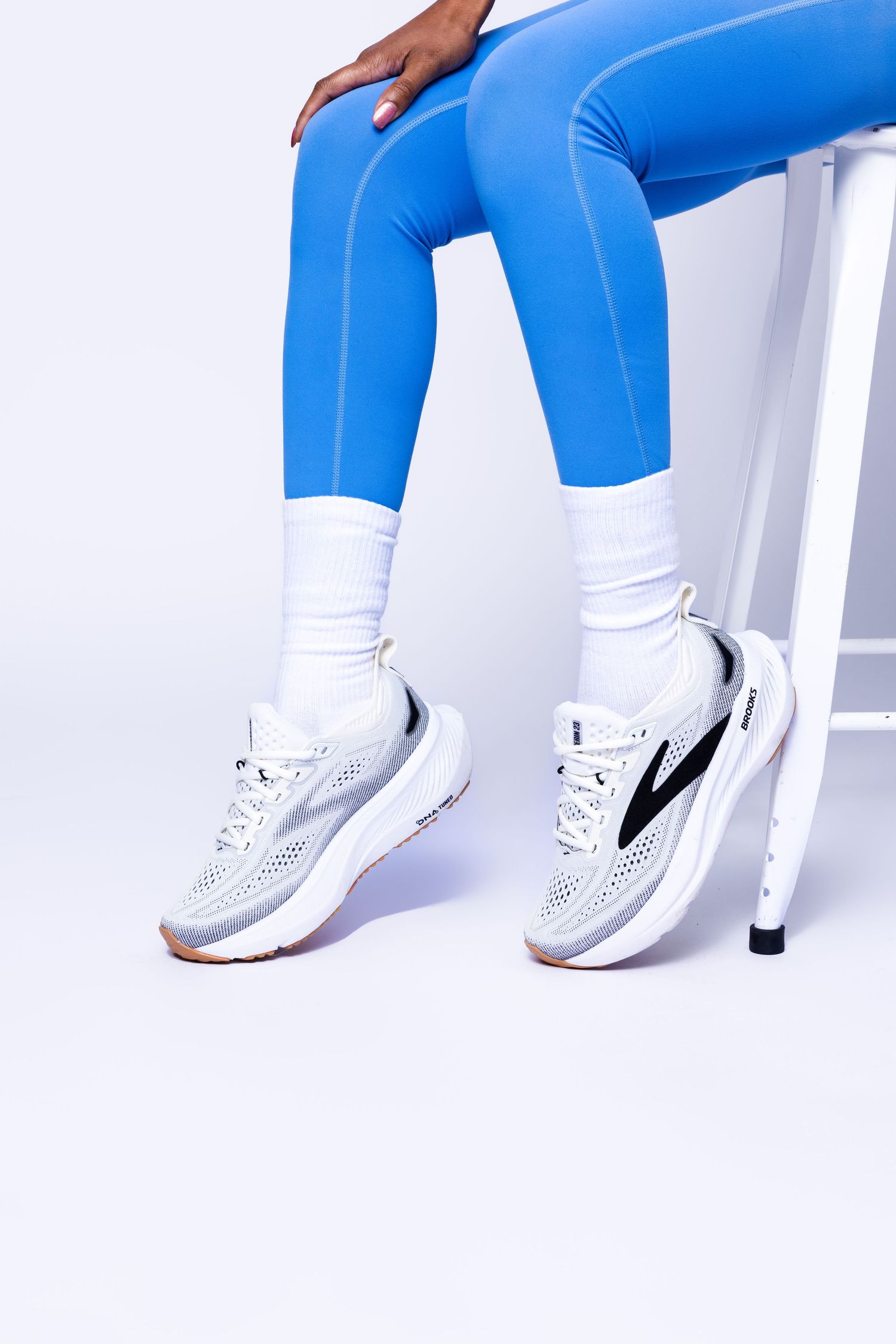 Person wearing blue leggings, white socks, and white Brooks running sneakers seated on a white stool.