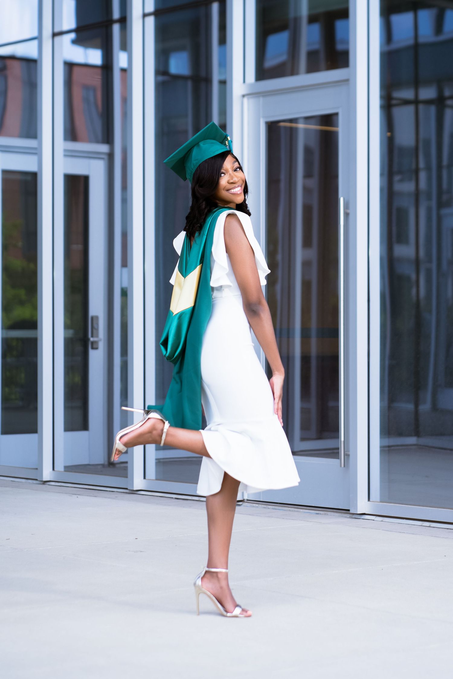 Dynamic graduation poses in white dress and green cap against modern glass architecture.