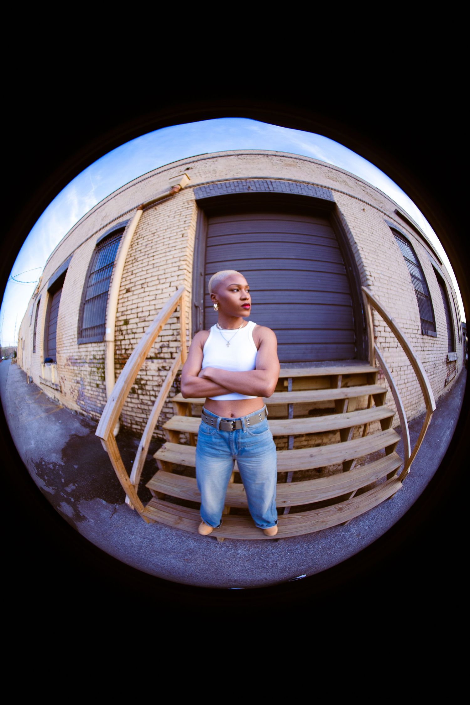 Fisheye view of person in white tank top and jeans standing on wooden steps near brick wall.