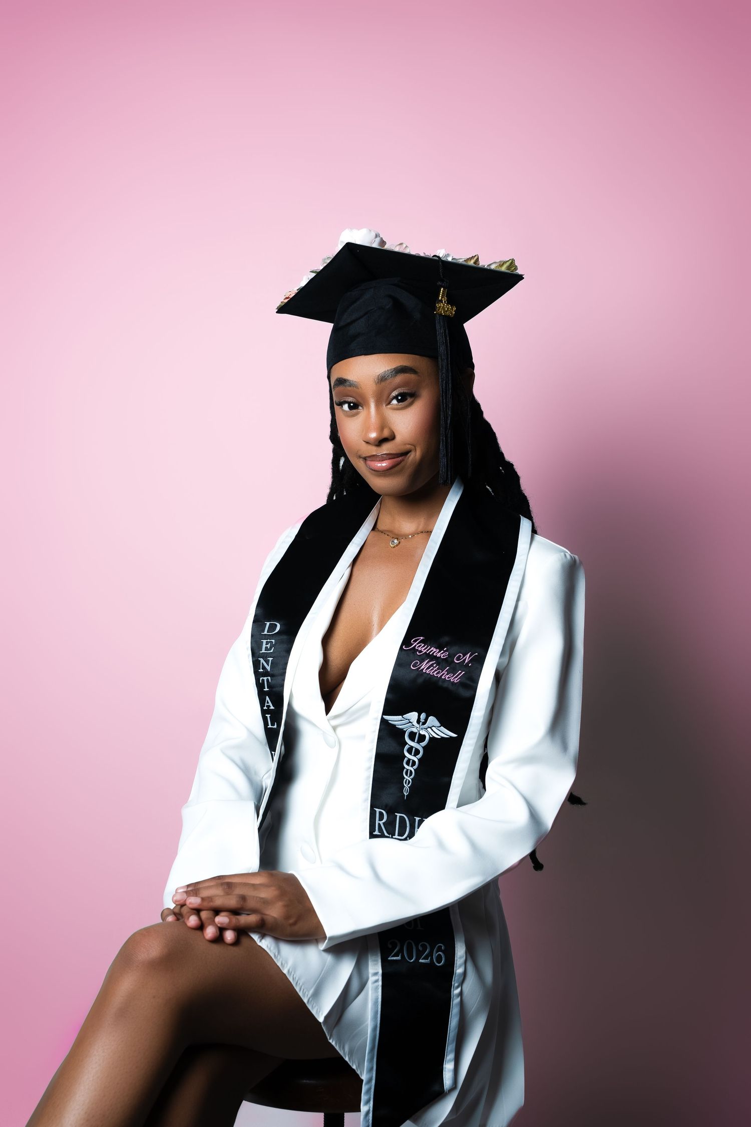 Graduate wearing black and white graduation stole and cap poses against pink background in studio portrait.