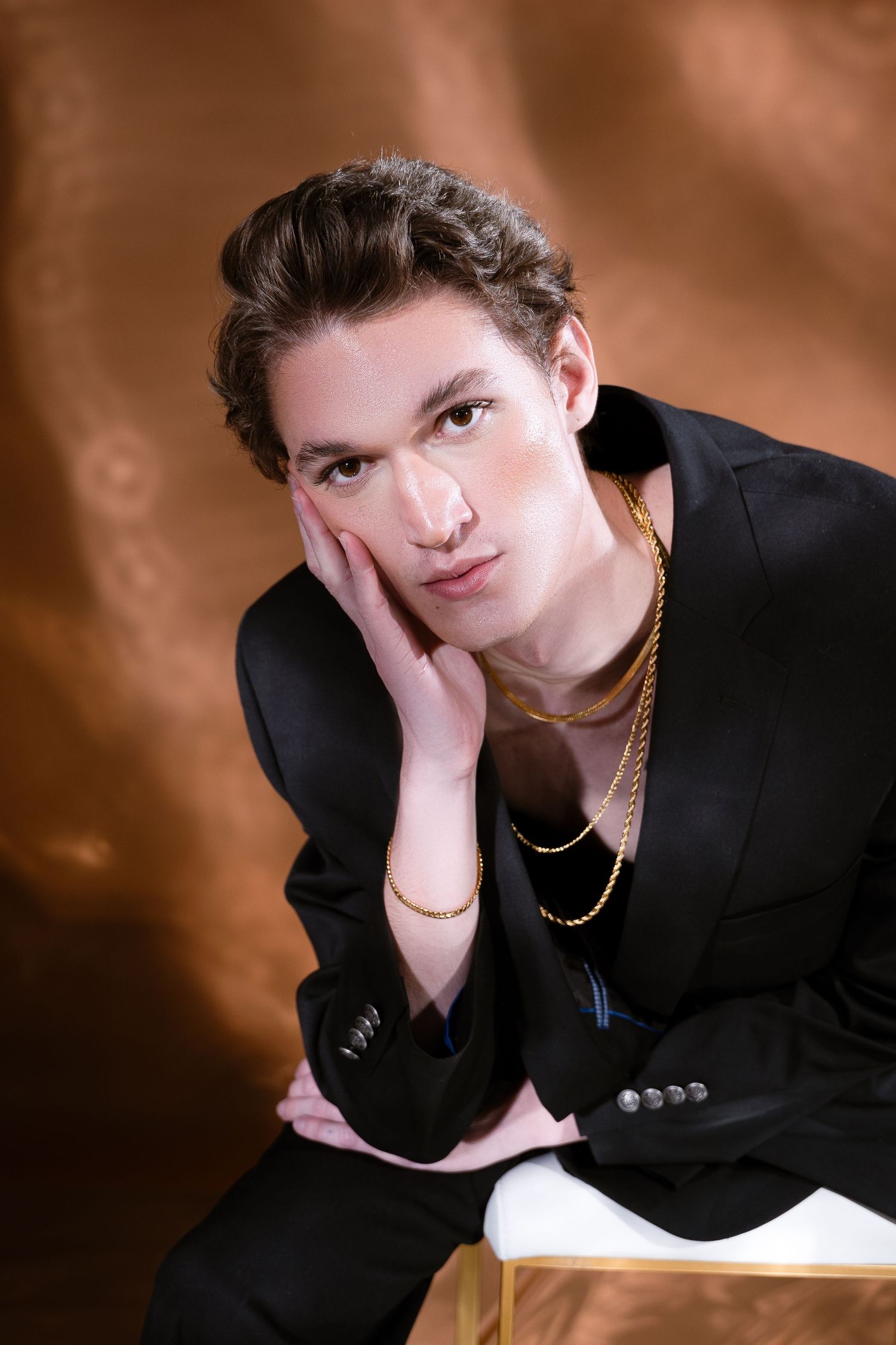 Stylish portrait in black blazer with gold necklaces against brown backdrop.