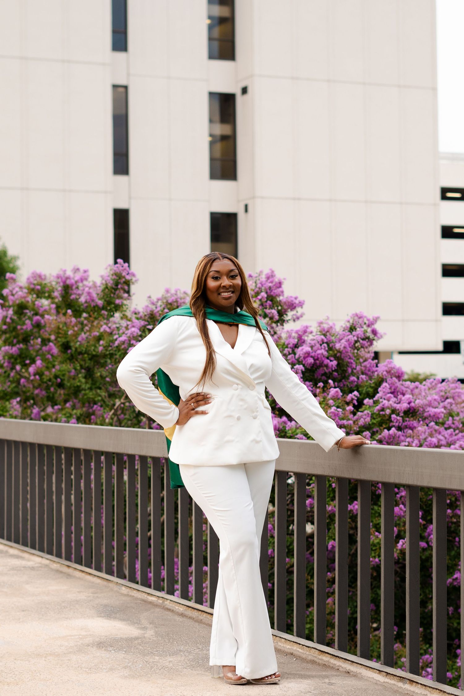 Graduate in white outfit posing near flowering bushes outside building.