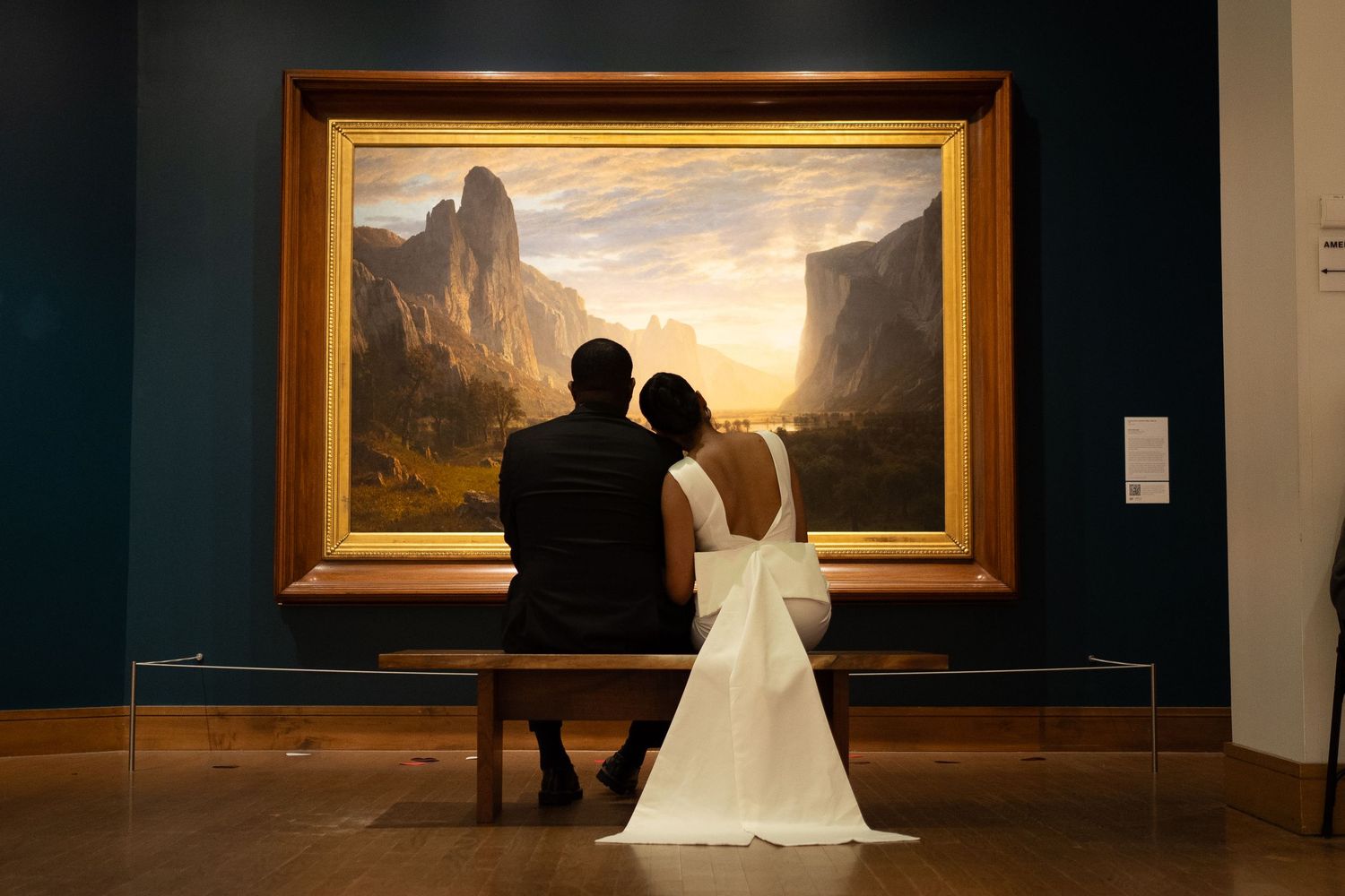 Newlywed couple in wedding attire sitting on bench admiring large landscape painting in art museum gallery.