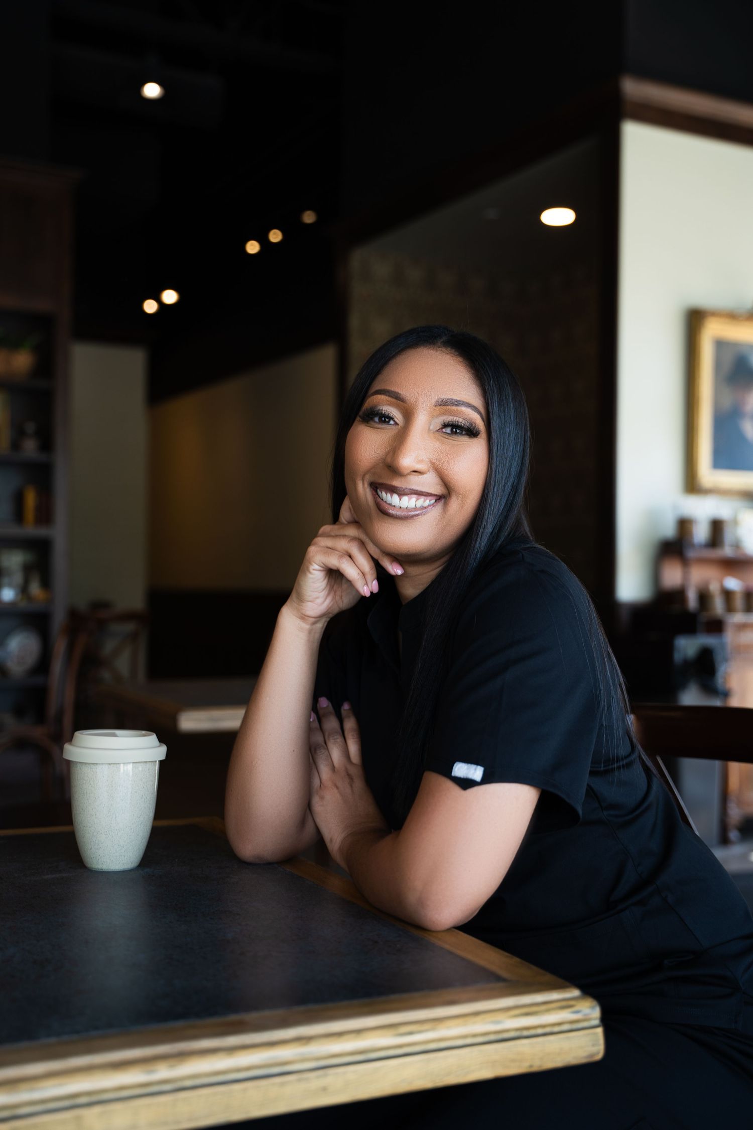 A woman wearing a black hijab sits at a cafe table smiling warmly while resting her head on her hand.