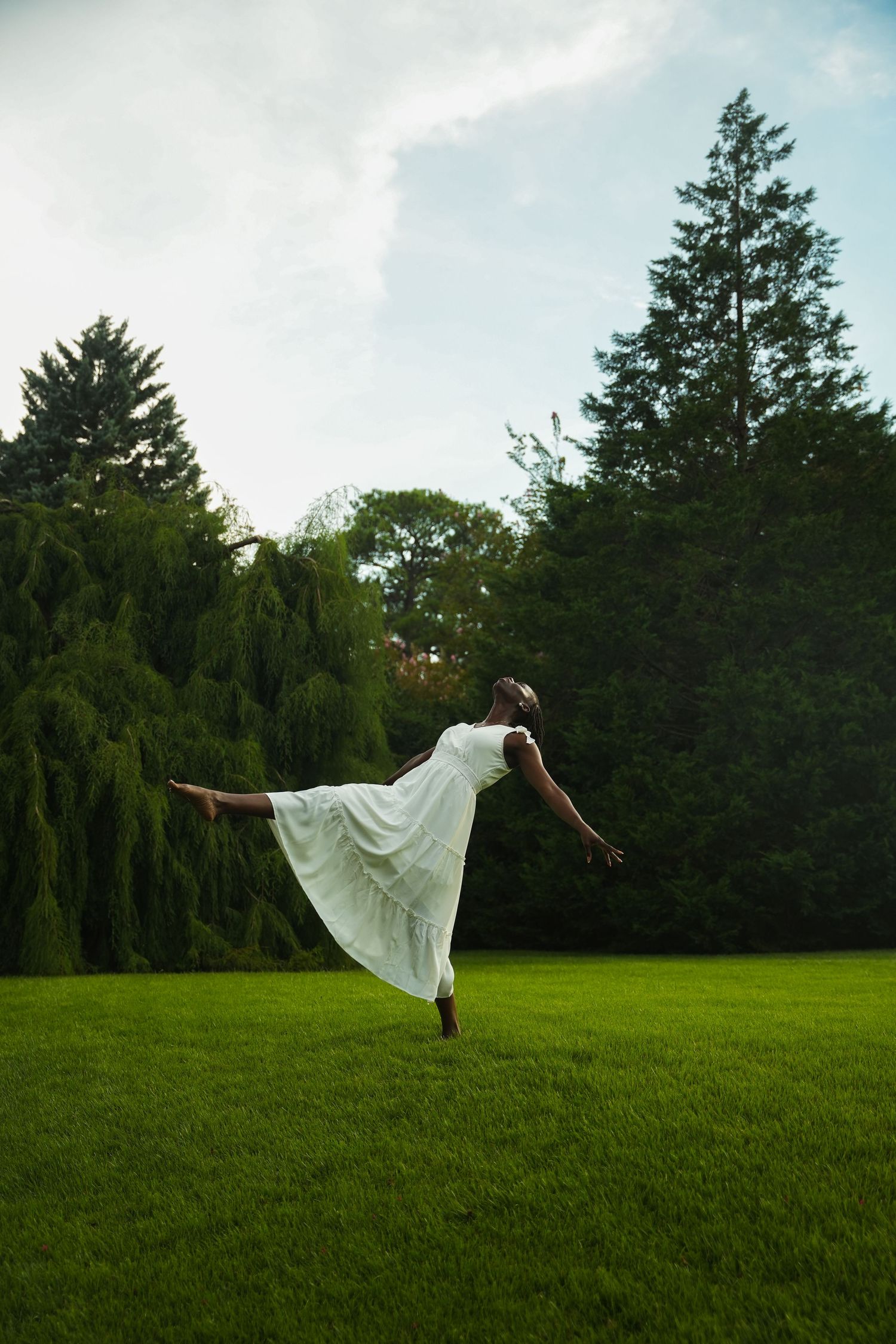 A person in a flowing white dress performs a graceful dance pose on a green lawn with trees in the background.