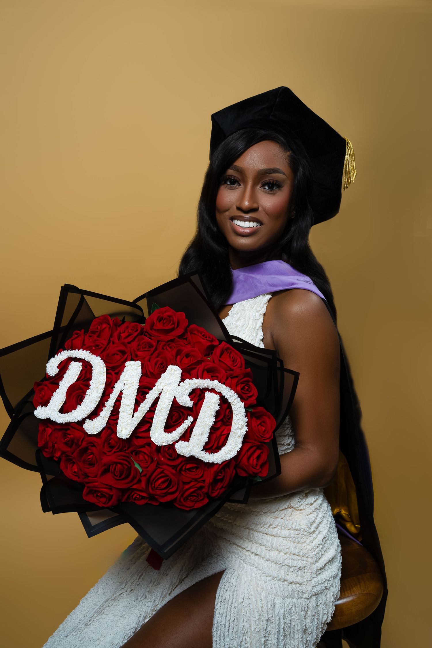 Graduate in white dress holds decorative red rose arrangement with DMD letters against golden background.