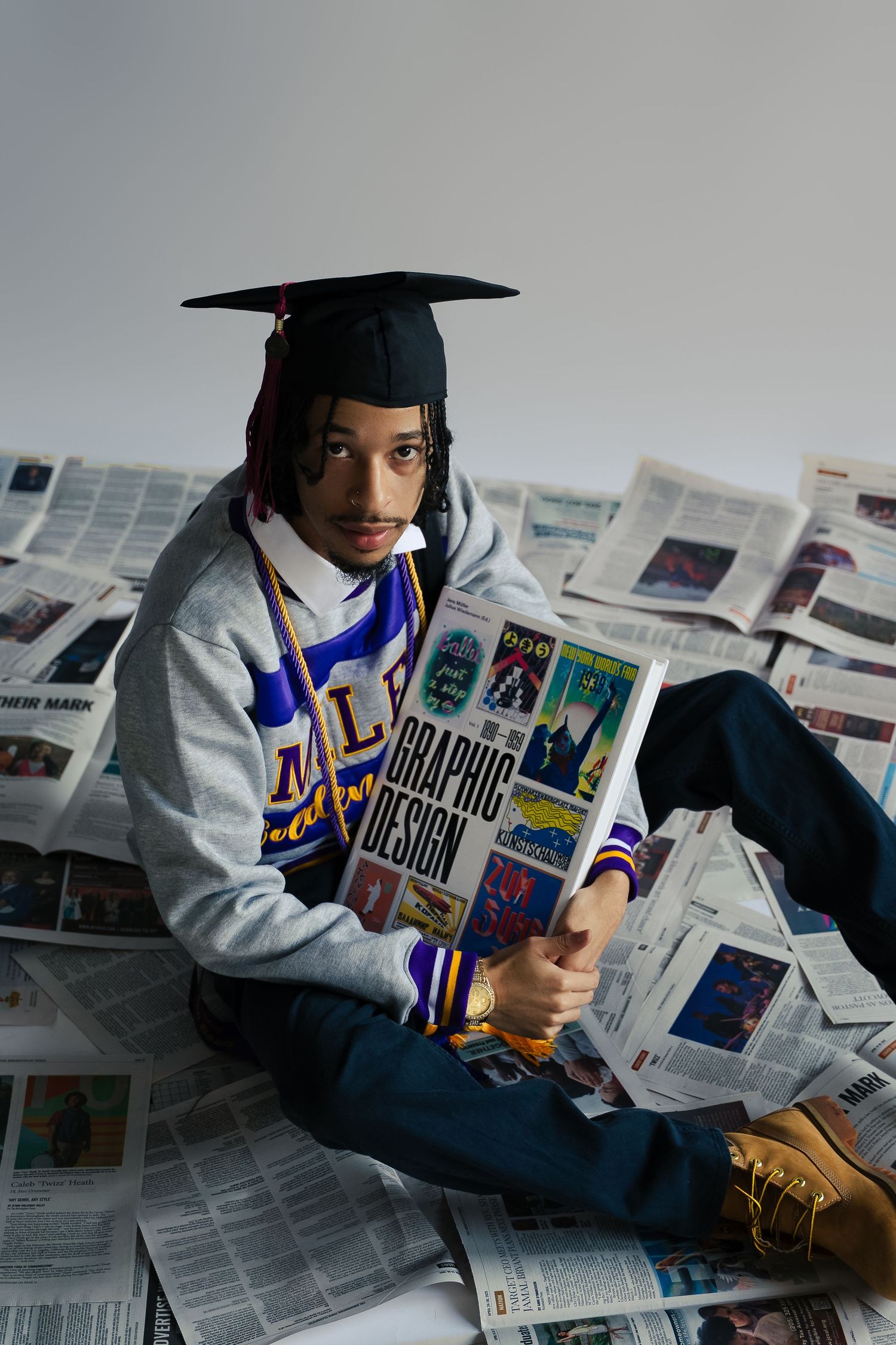 Graduate in casual attire sits surrounded by newspaper clippings.