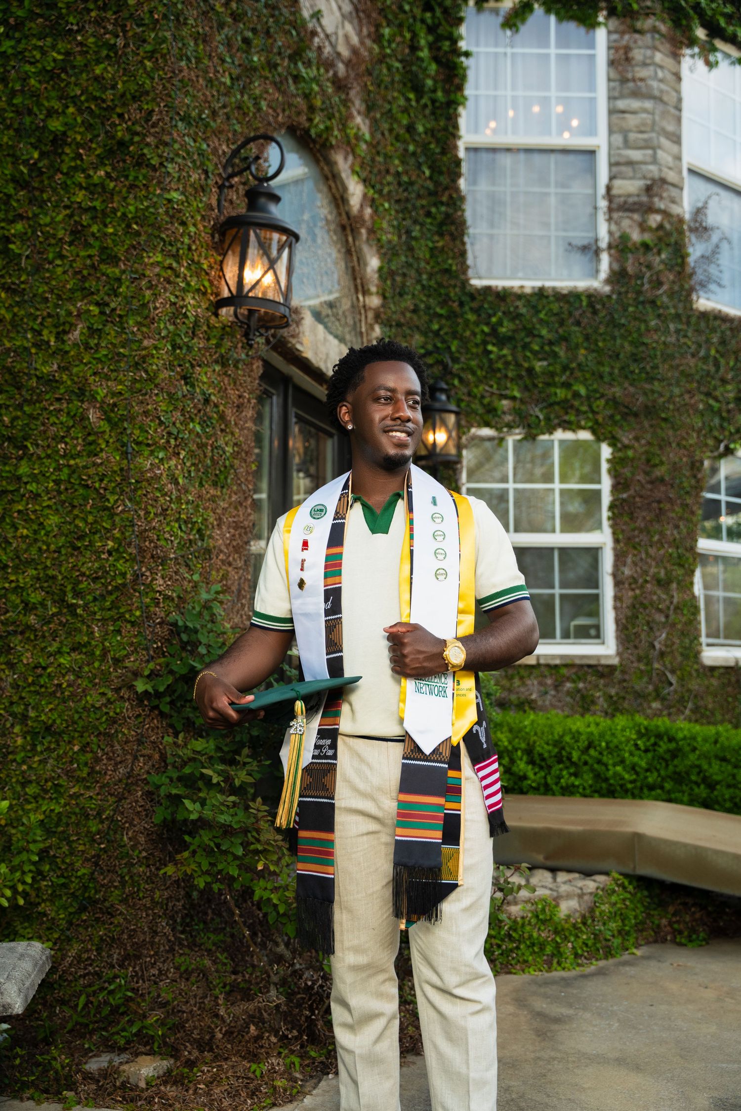 Smiling graduate in kente stole and academic regalia stands before ivy-covered building during outdoor graduation photo.
