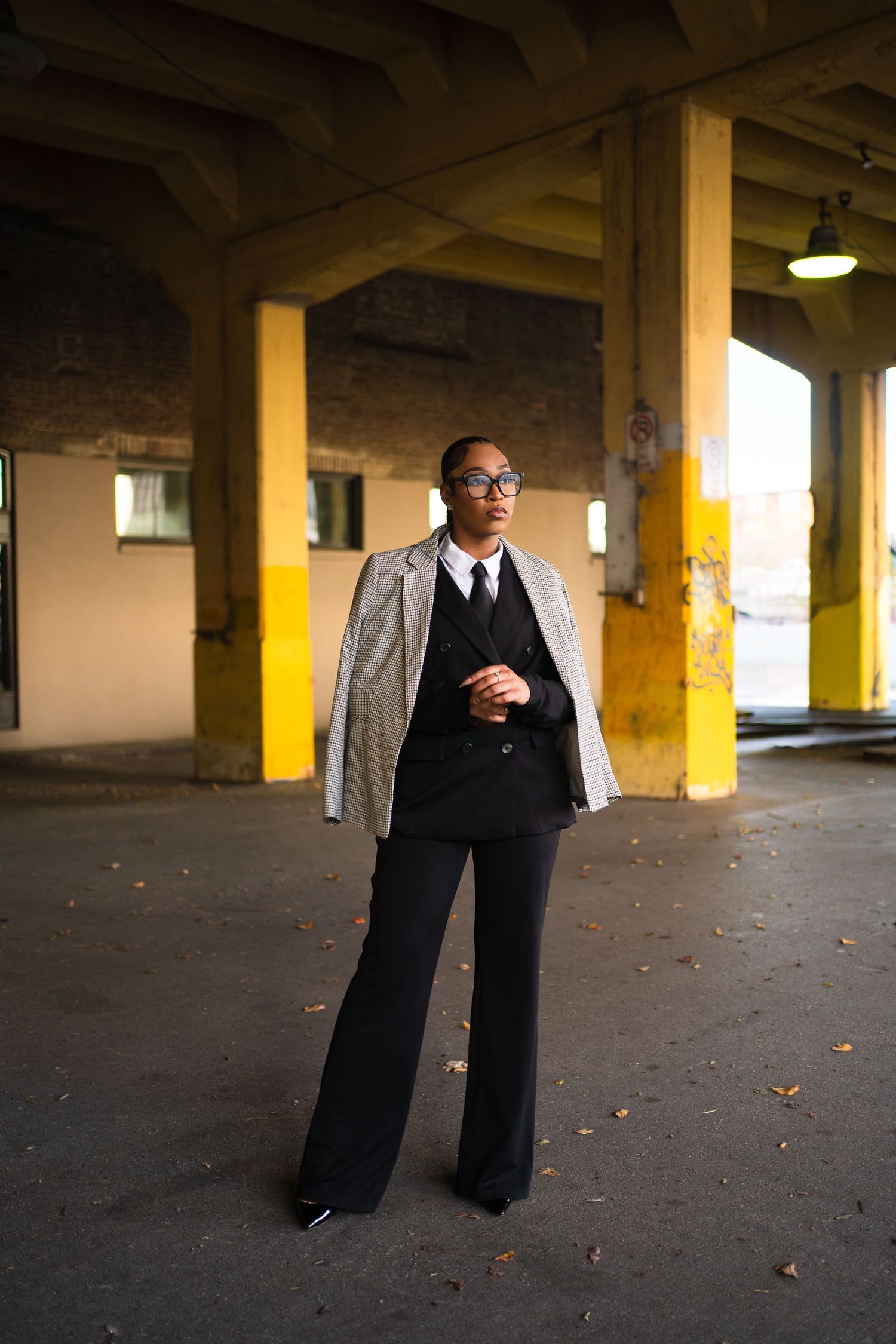 Person in formal attire sits contemplatively in a dimly lit concrete parking garage with yellow pillars.