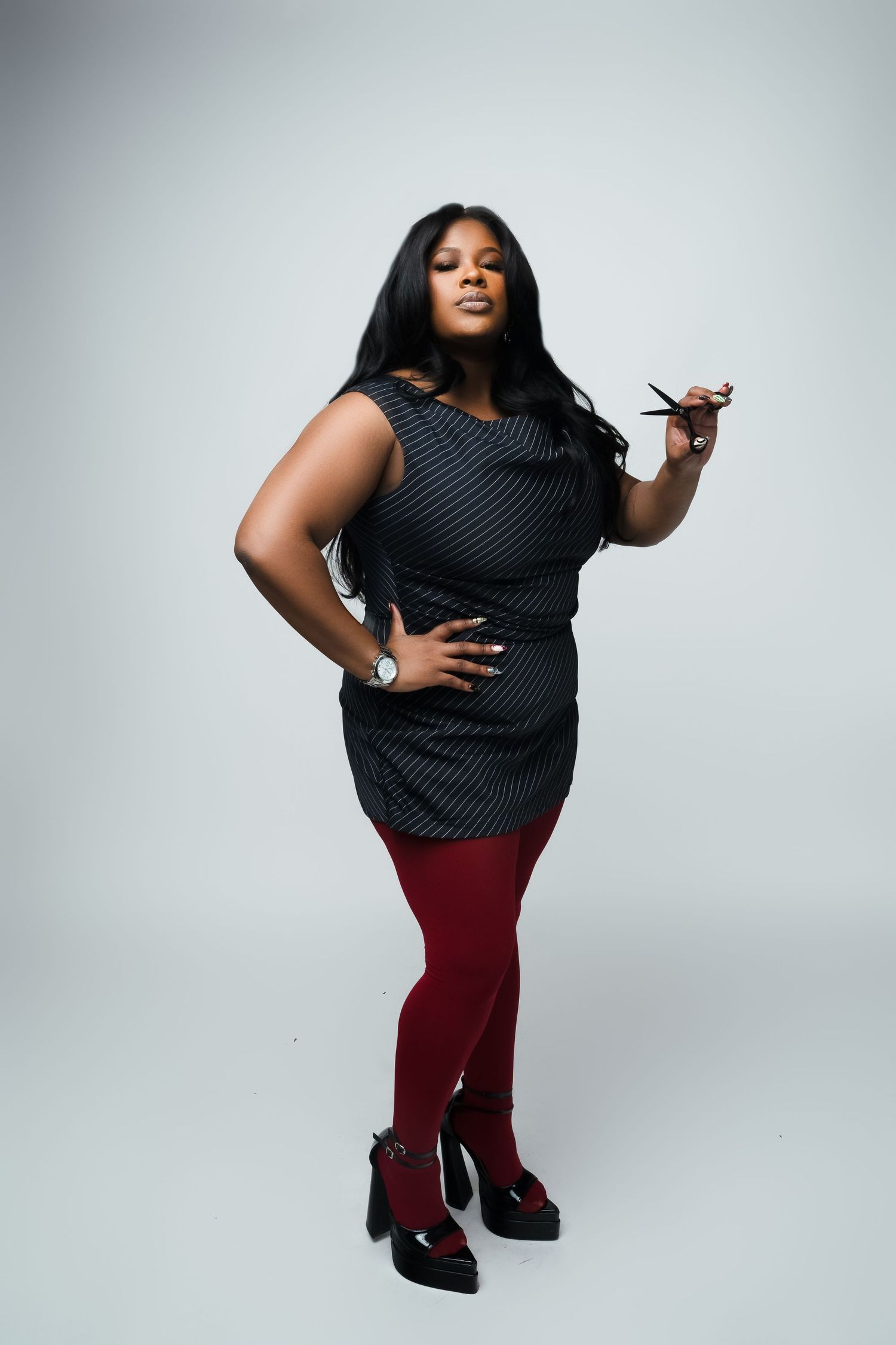 A plus-size woman poses confidently in a black dress, red tights, and heels, holding scissors against a gray background.