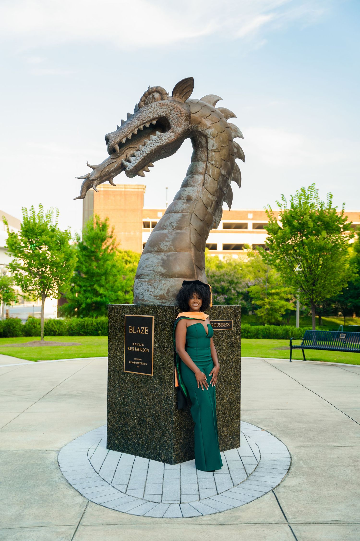 A dragon sculpture made of metal rises from a granite base in a public plaza surrounded by trees and buildings.