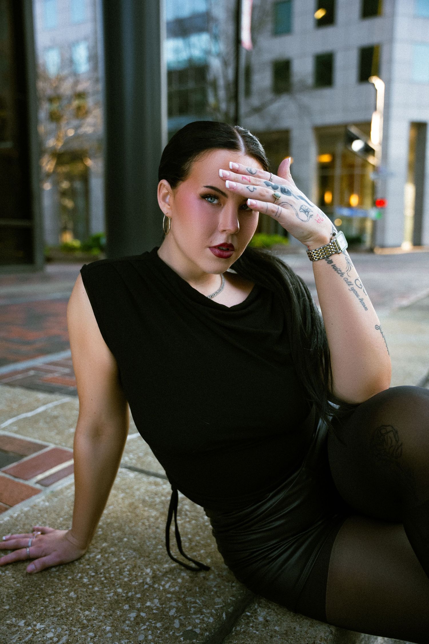 A person in a black outfit poses dramatically on a brick surface in an urban setting.