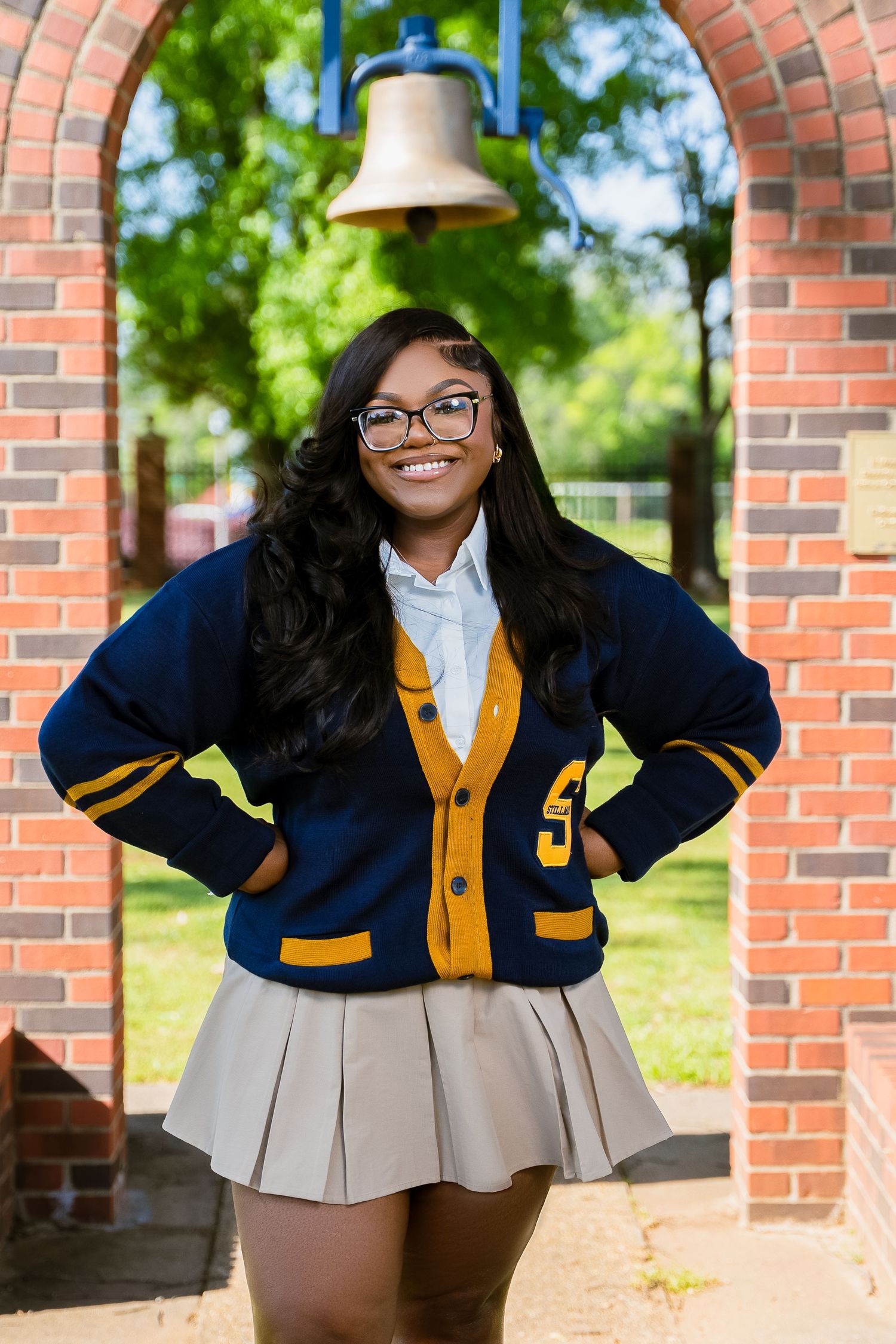 Student in navy and gold varsity sweater poses under brick archway with bell.