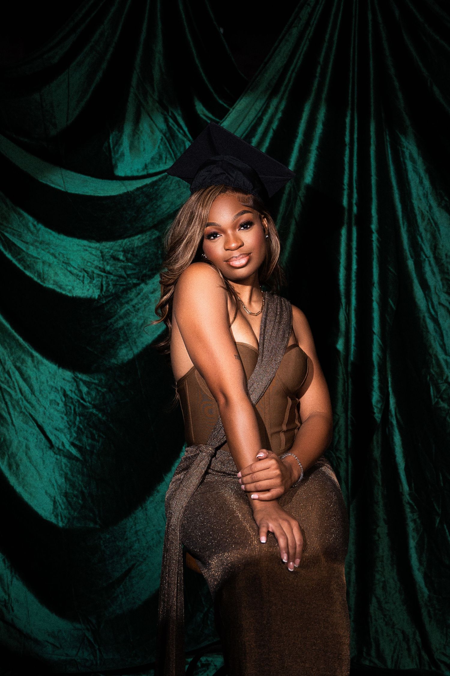 Glamour portrait against dark emerald green velvet backdrop featuring elegant brown dress and dramatic lighting.