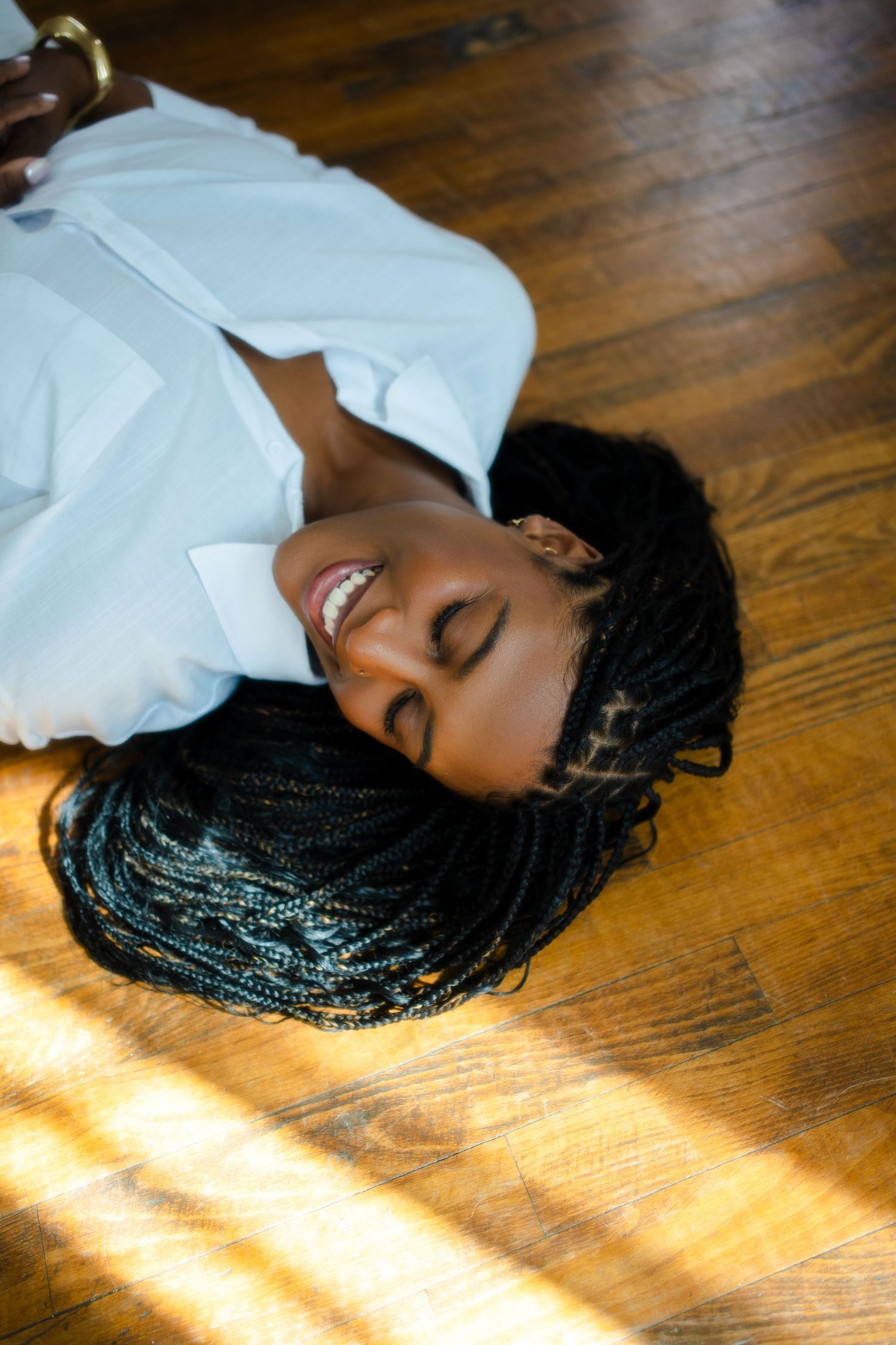A person with braided hair lies peacefully on a wooden floor as sunlight streams through nearby windows.