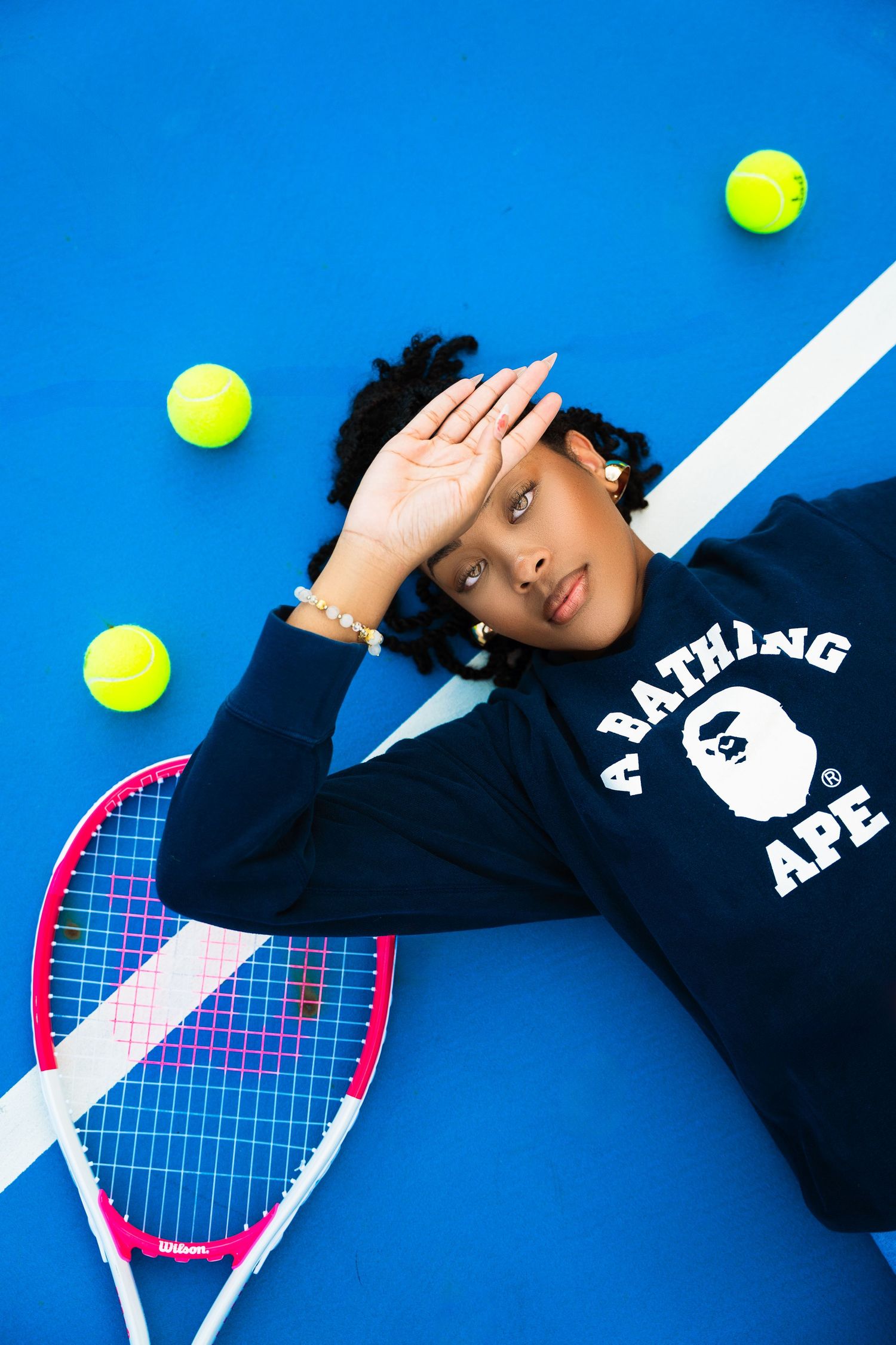 Tennis player in navy sweatshirt lying on blue surface with tennis balls and pink racket in creative composition.