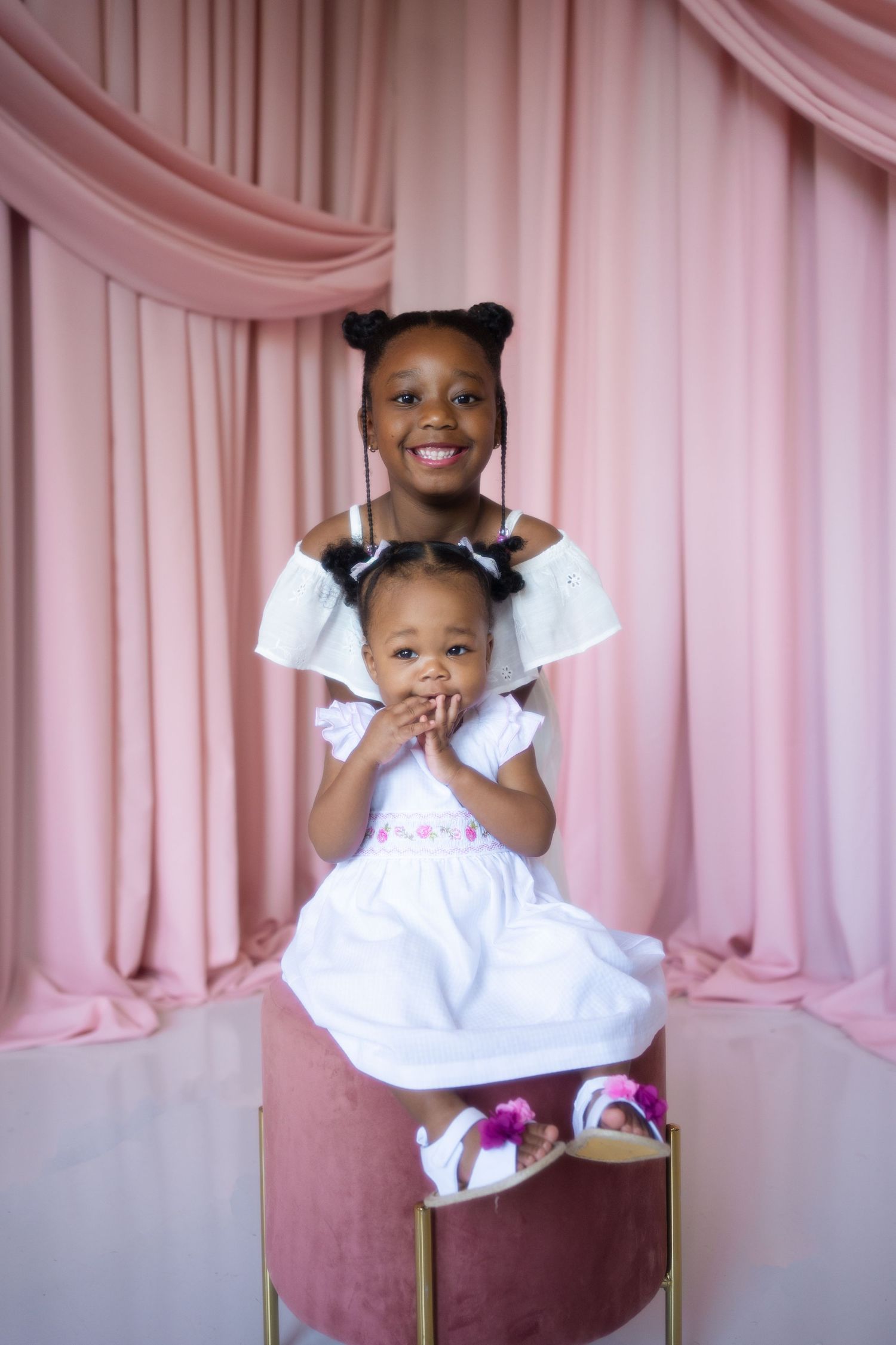 Two young girls in white dresses pose together in front of pink drapes for a professional portrait photo.