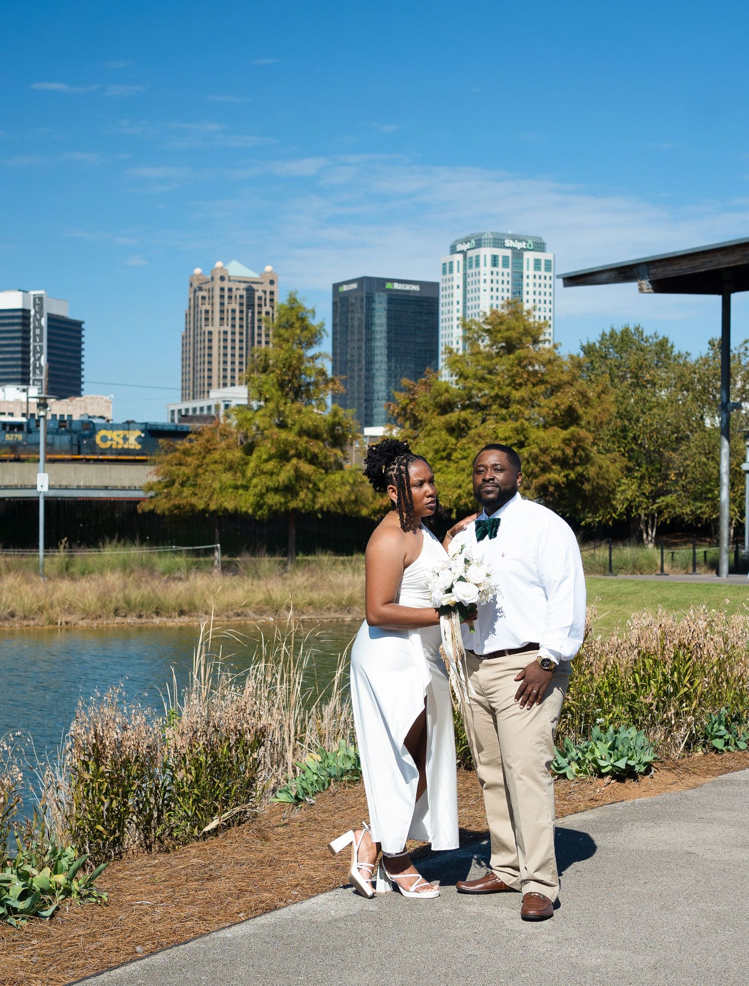 A couple poses together in elegant attire by a scenic waterfront with city buildings in the background.