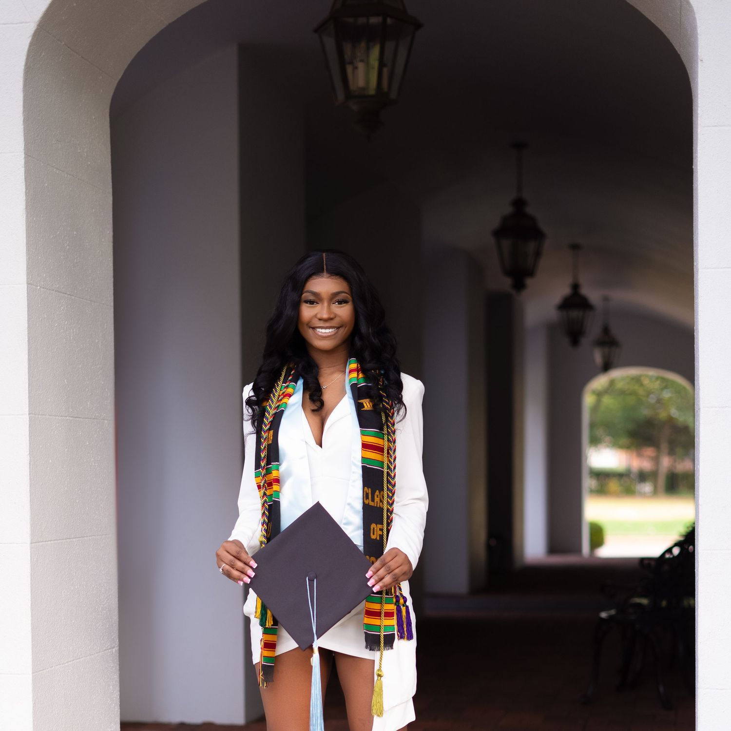 Graduate in white vest and graduation regalia poses under arched walkway with lanterns.
