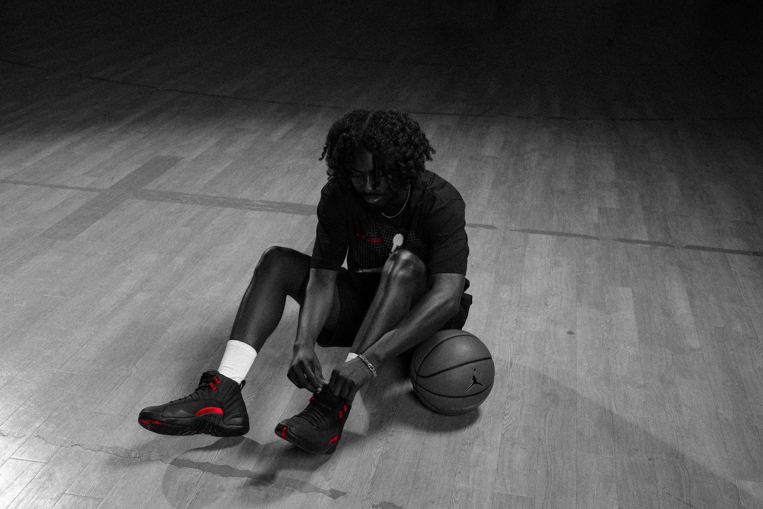 Basketball player sitting on court lacing up red and black sneakers with a basketball beside him.