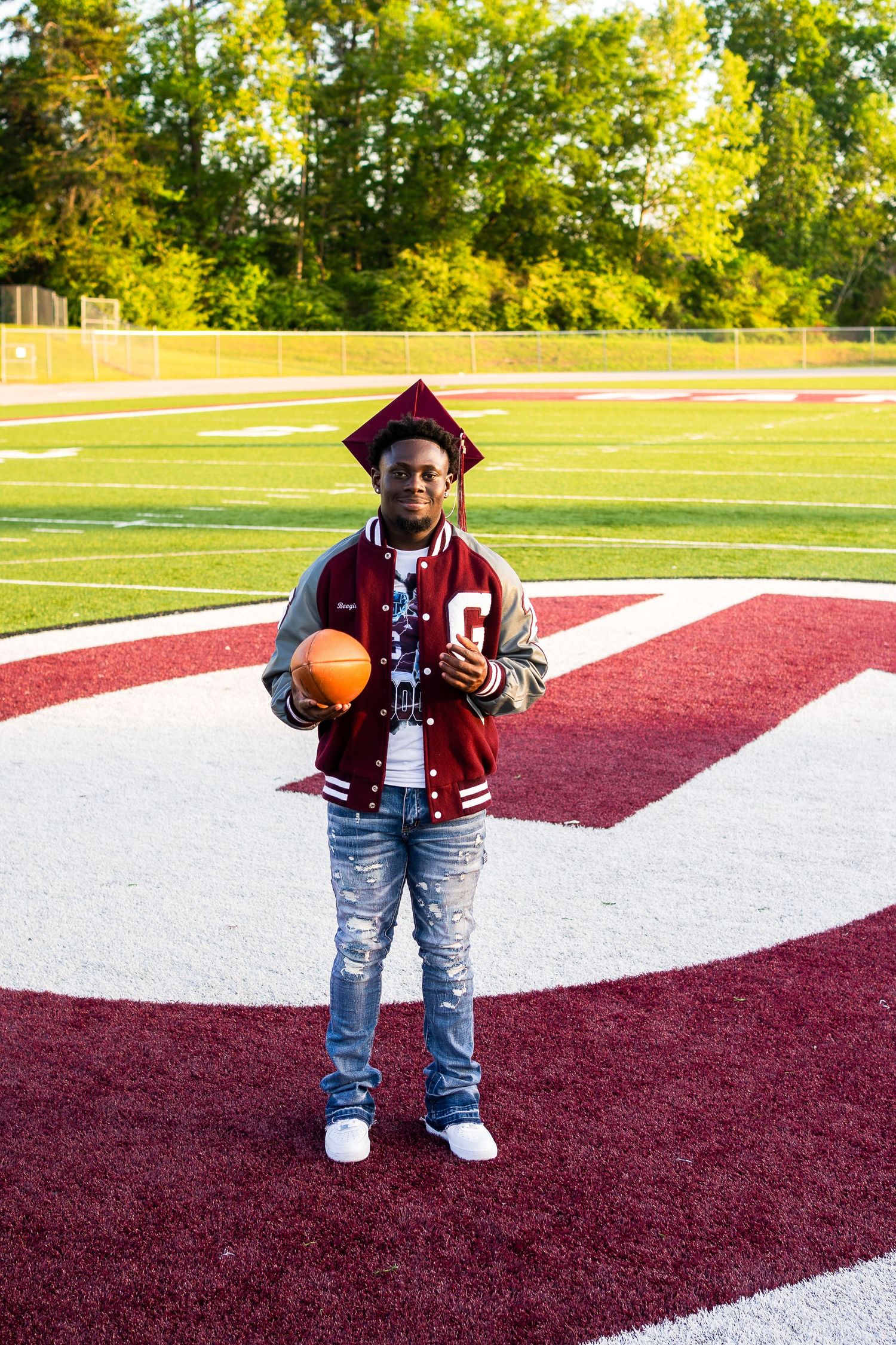 Graduate in varsity jacket posing with basketball on red field markings.