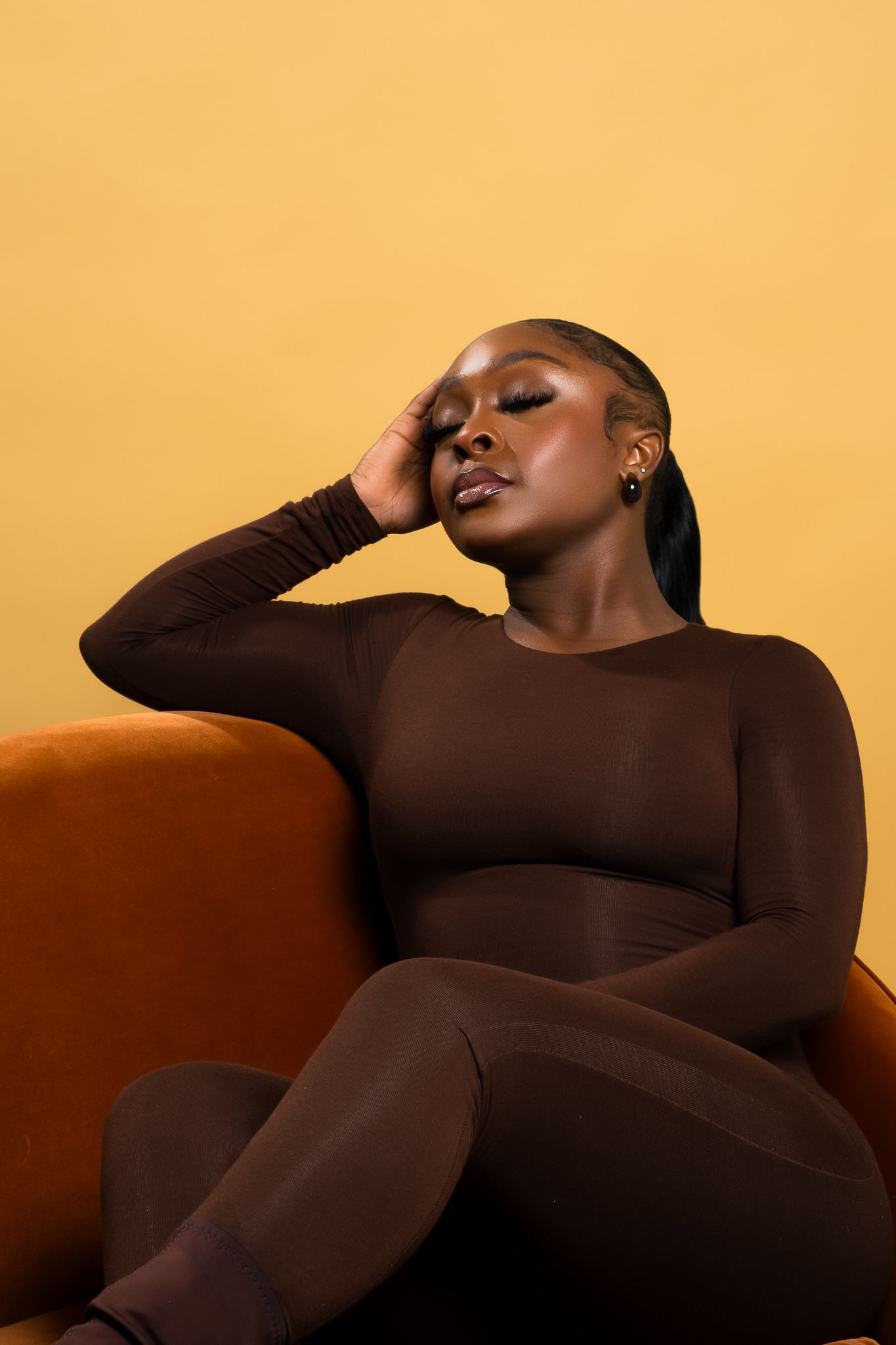 Fashion model in brown fitted outfit poses on orange velvet chair.