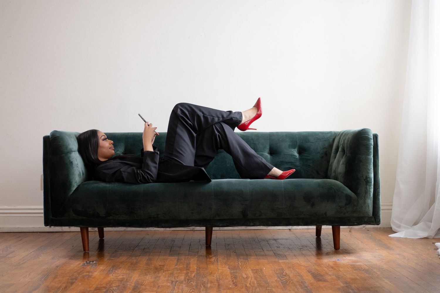 Person in black attire lying on a green velvet couch wearing red heels.
