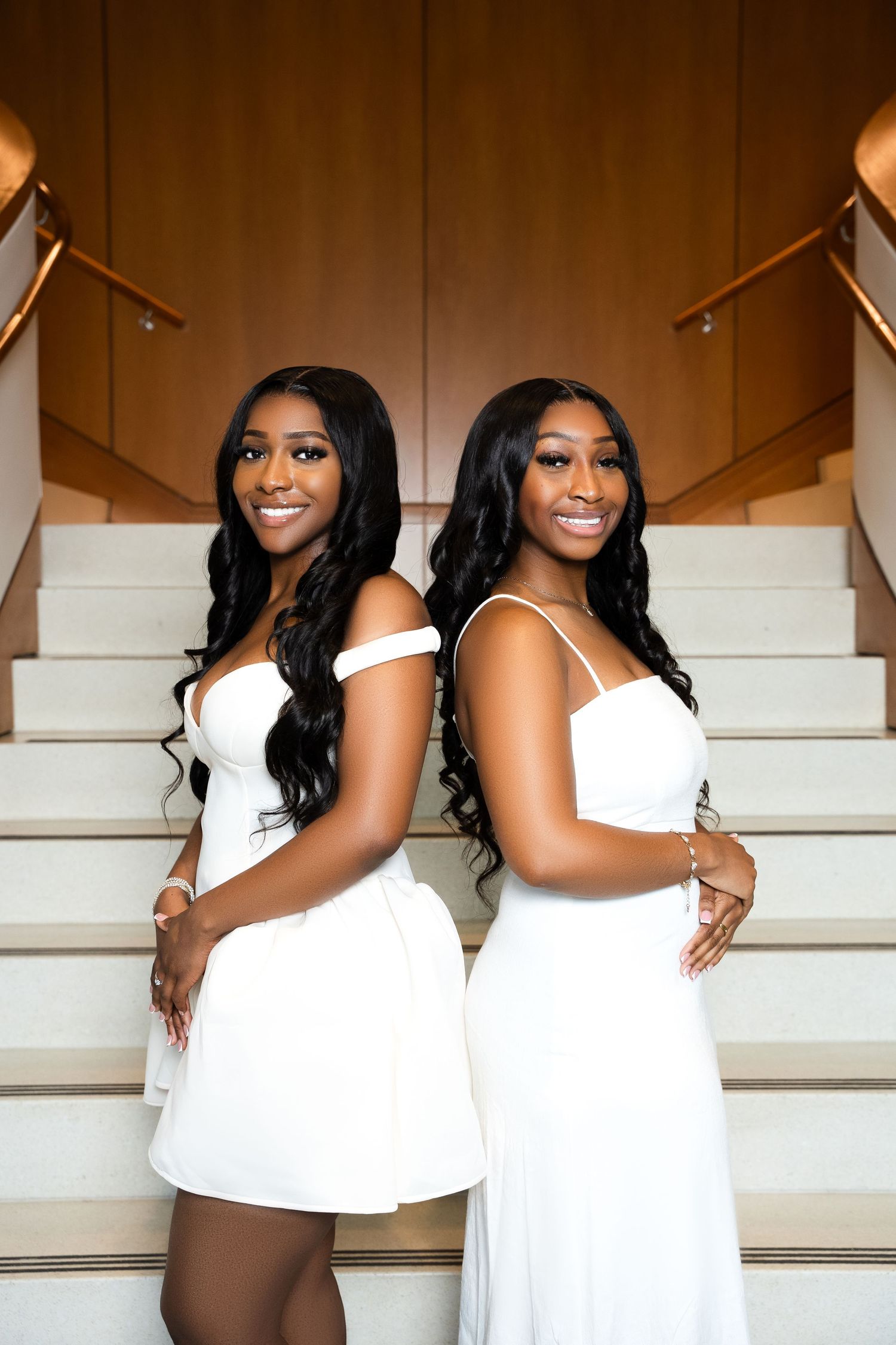 Two young women in elegant white dresses pose back-to-back on a grand staircase, smiling confidently.