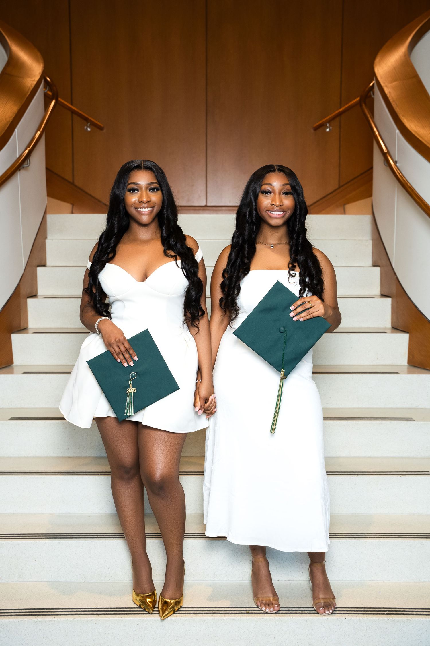 Two women in white dresses holding green graduation caps on stairs, celebrating their graduation day.