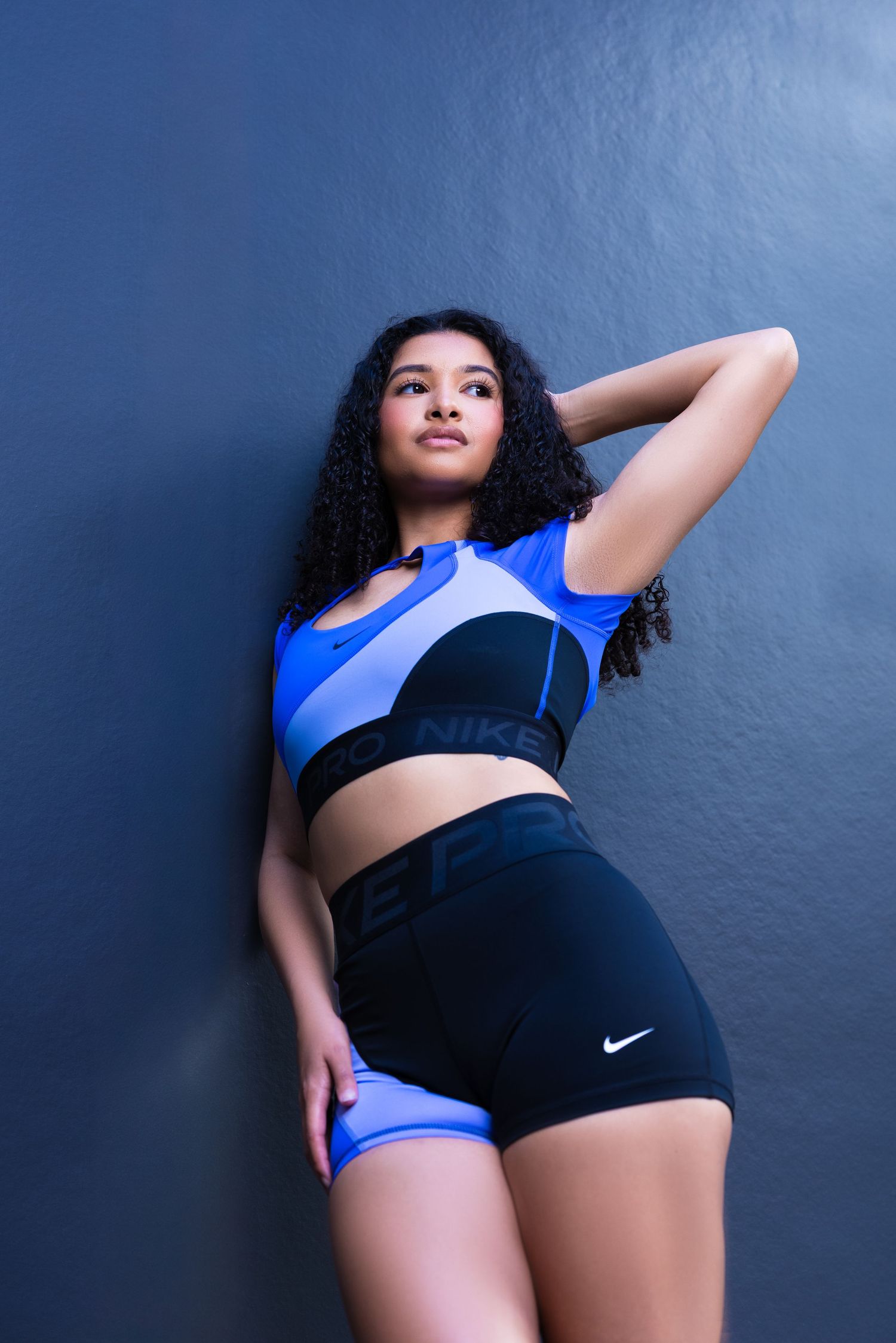Athletic figure in black Nike shorts and blue sports bra poses against blue wall in dynamic fitness photoshoot series.