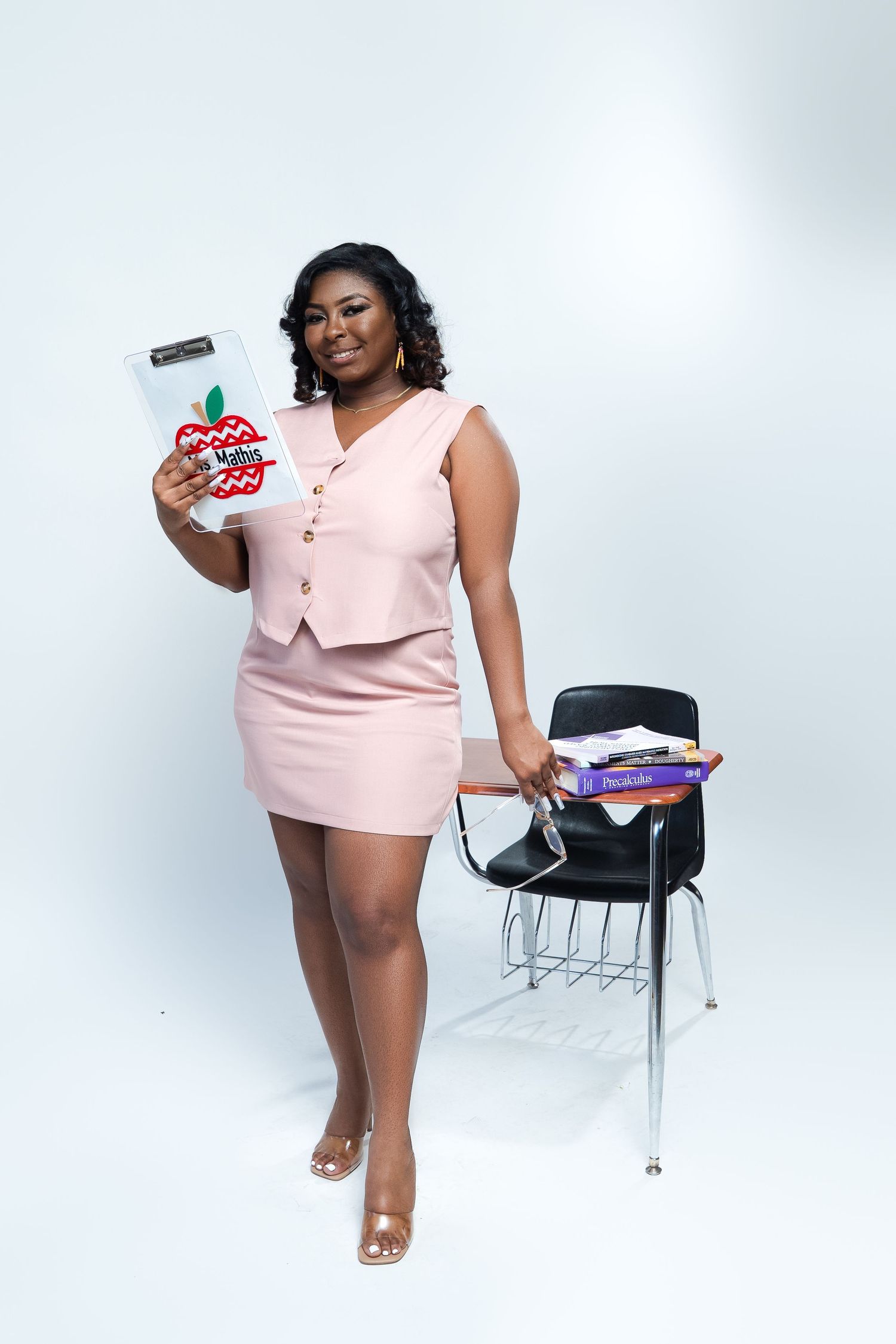 Professional in pink outfit poses with supplies near classroom chair.
