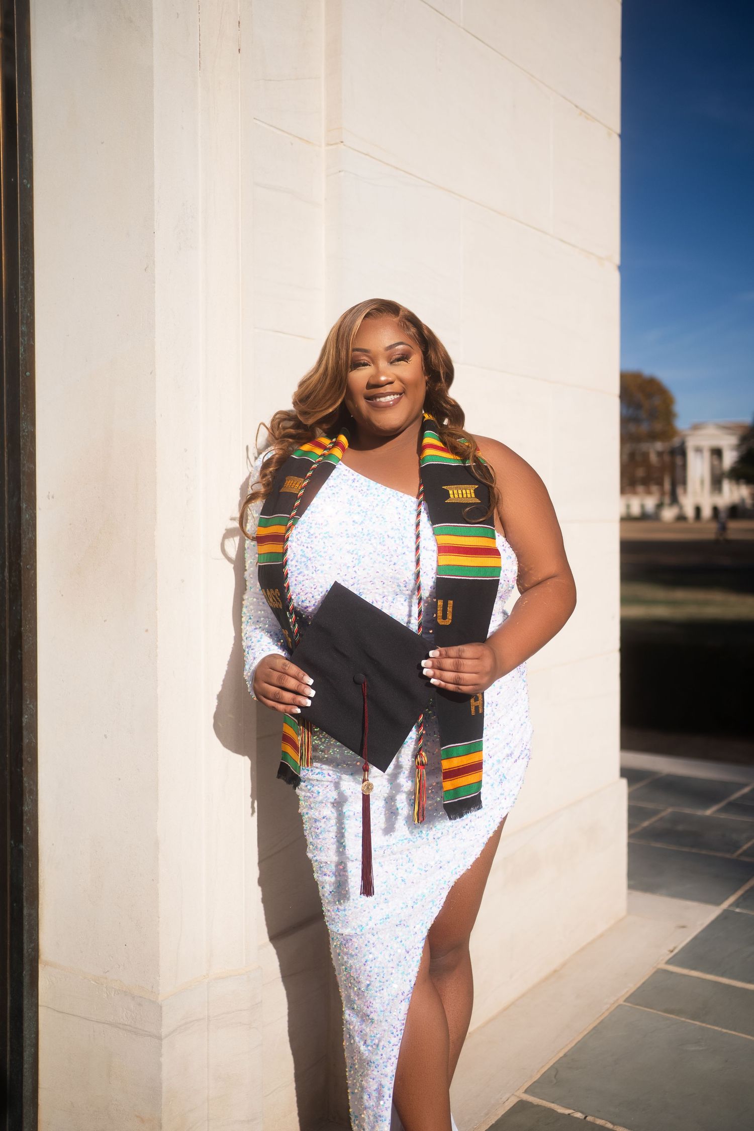 Graduate wears white dress with traditional kente stole against architectural backdrop.