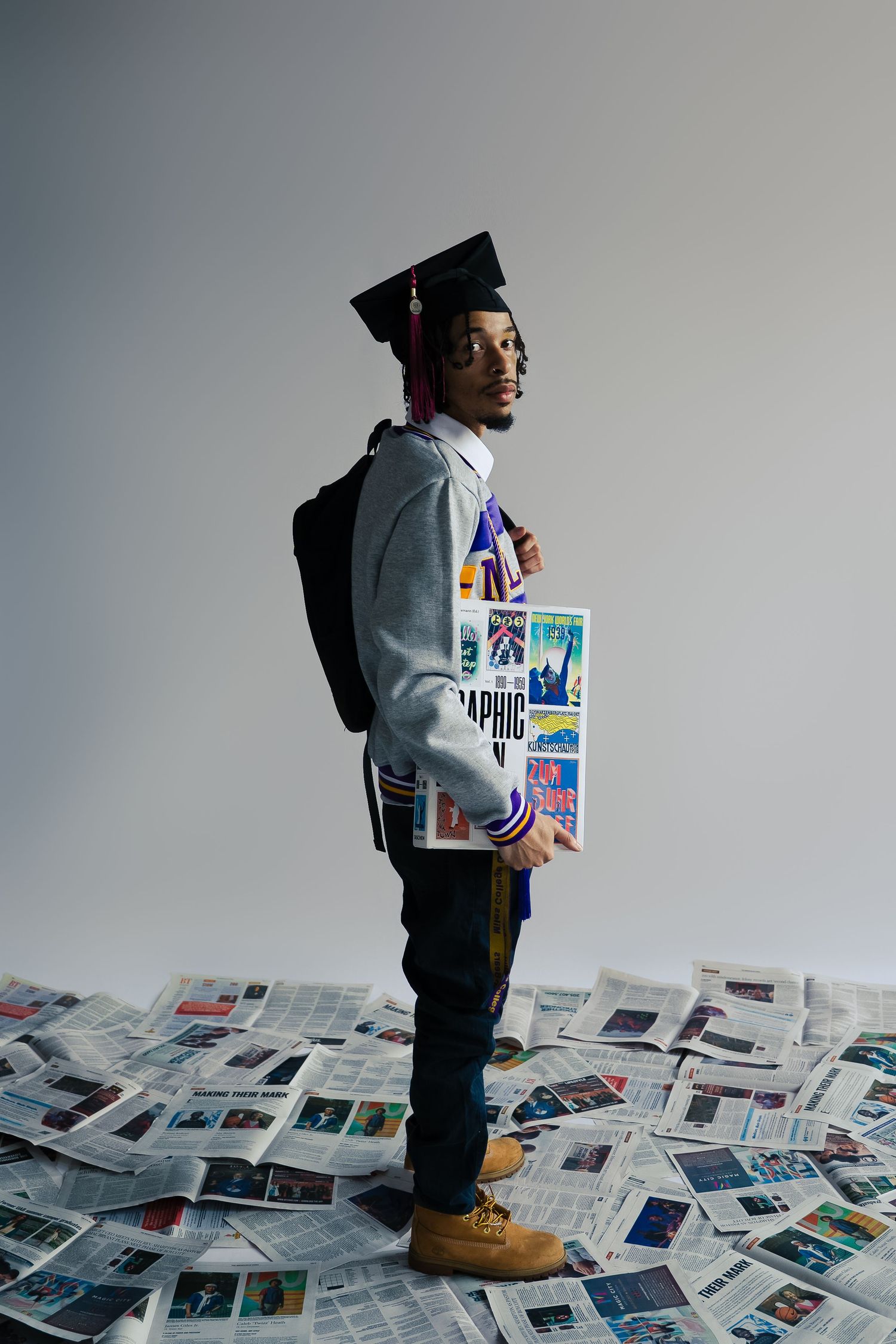 Graduate standing on scattered newspapers wearing a varsity jacket and graduation cap.