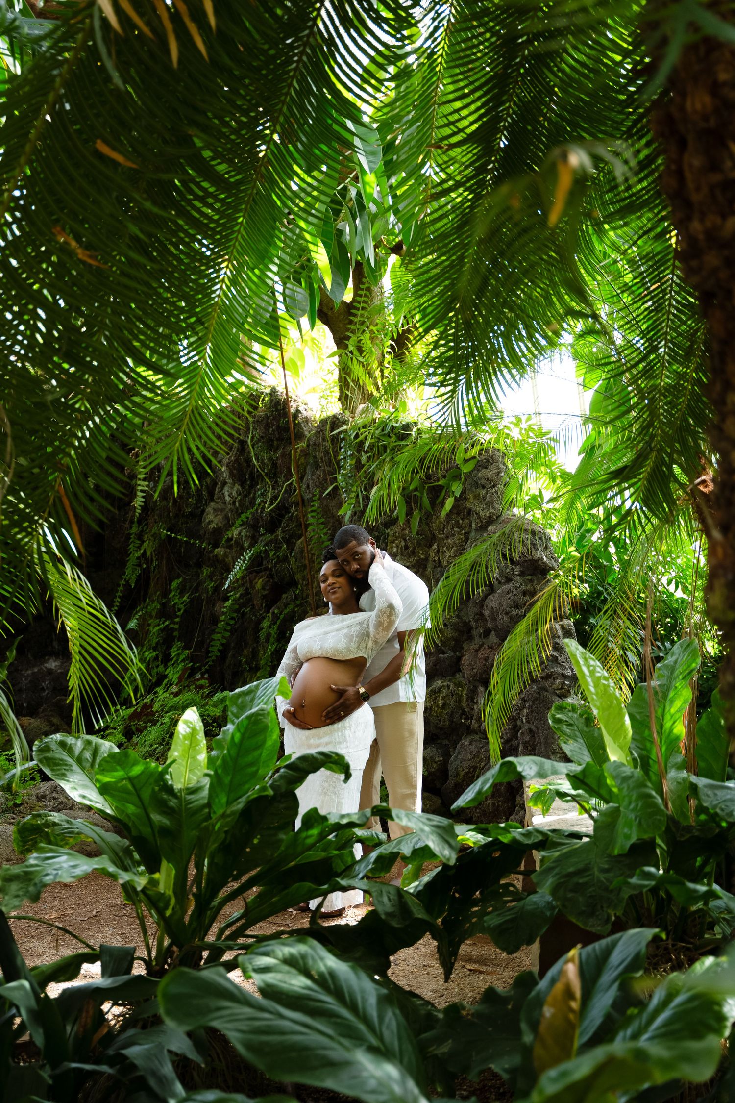 A romantic maternity photoshoot in a lush tropical garden setting with palm fronds.