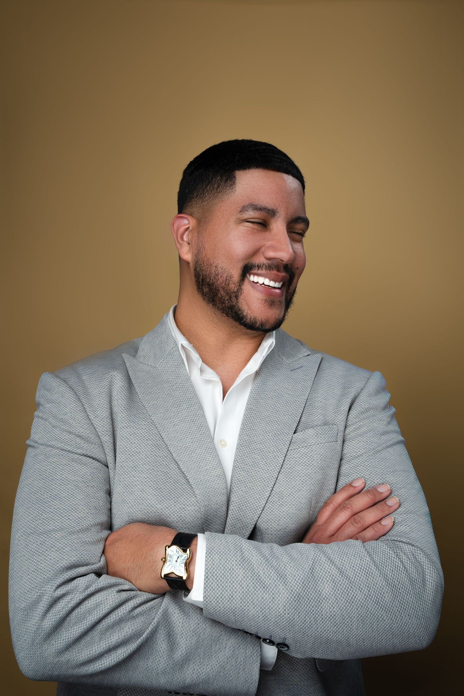 Smiling man in gray blazer and white shirt with arms crossed, wearing a watch, against a golden background.