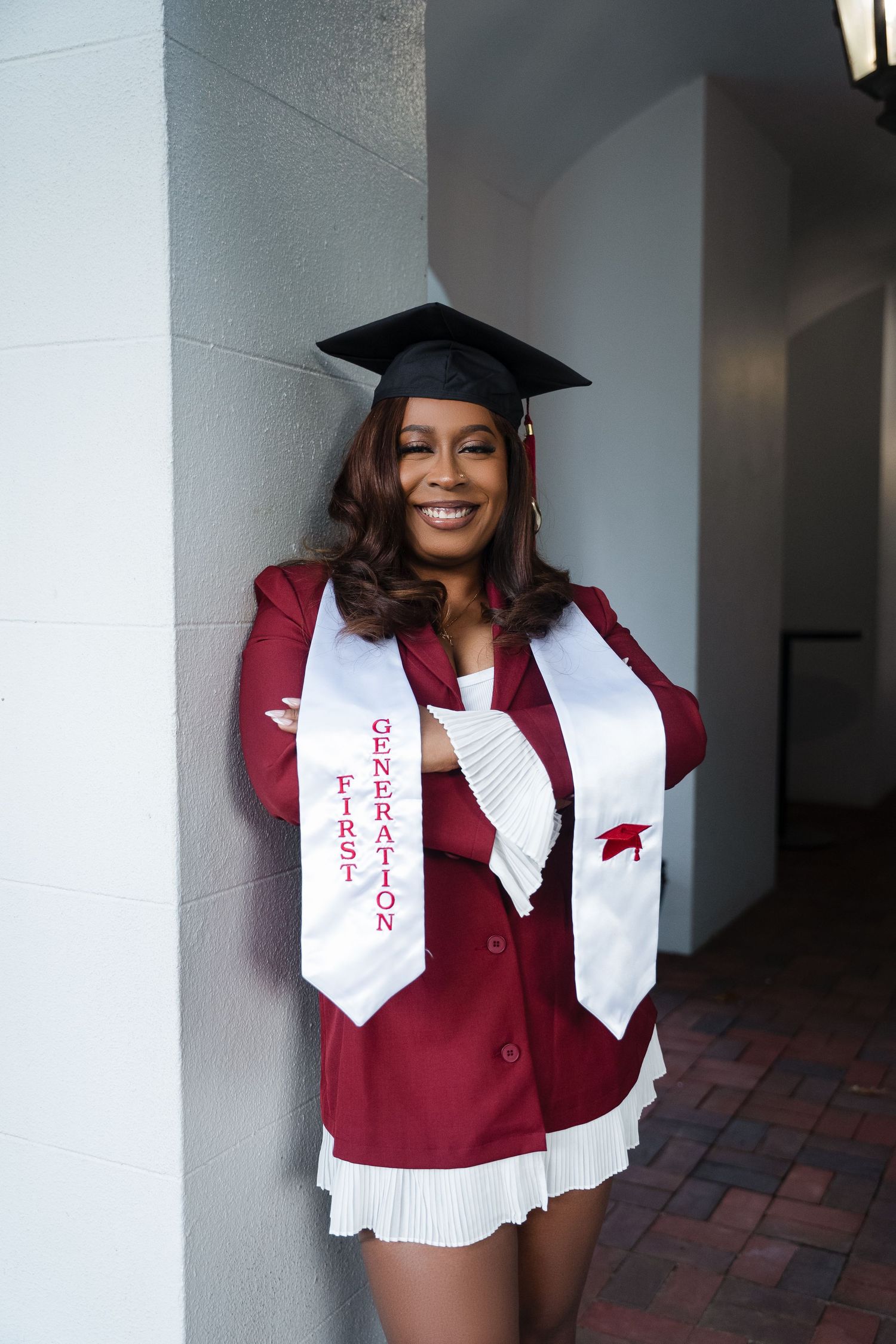 Graduate wearing a maroon blazer, white sash and graduation cap poses against a wall in celebration.