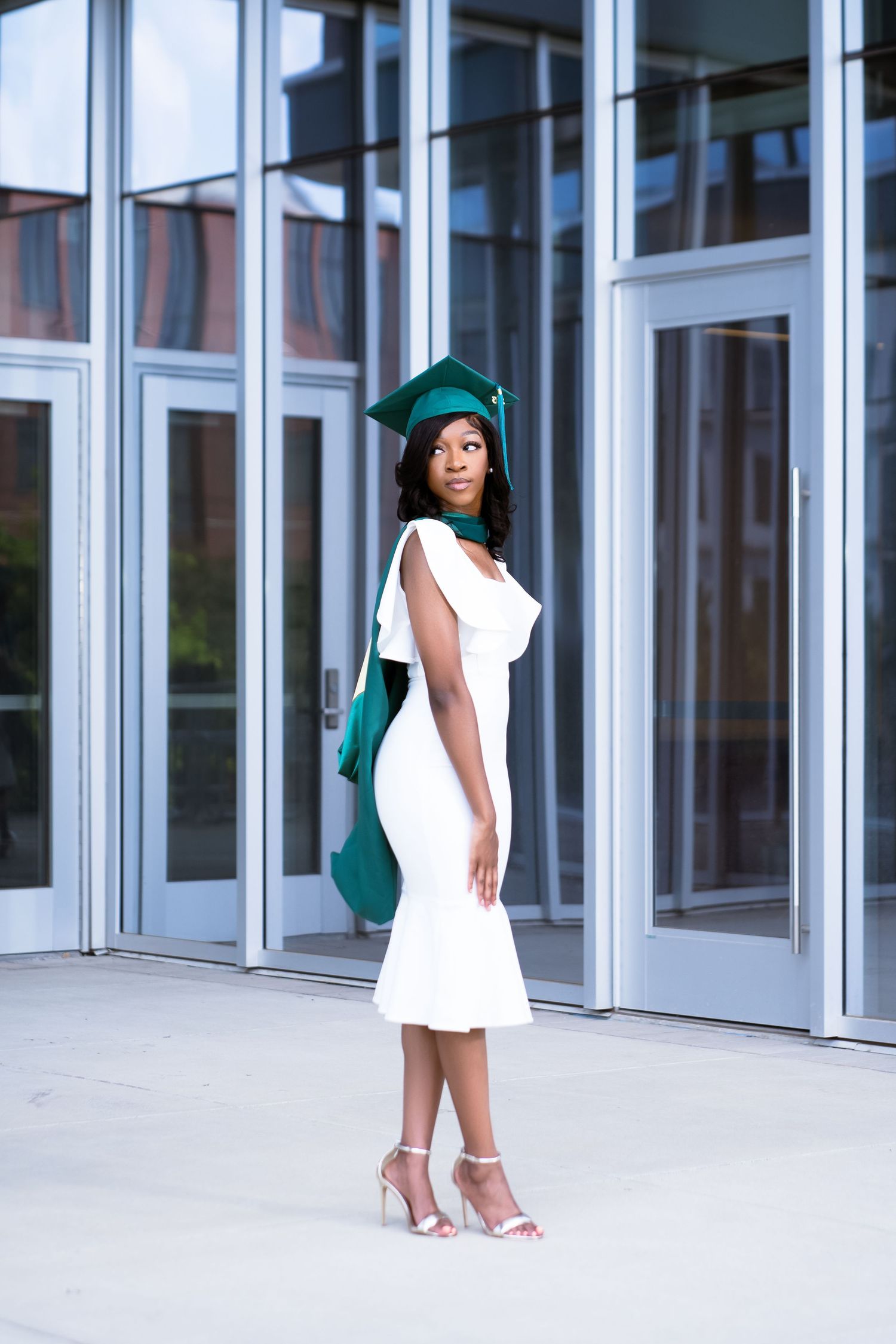 Elegant graduation photos featuring a white dress and emerald green cap against glass building.