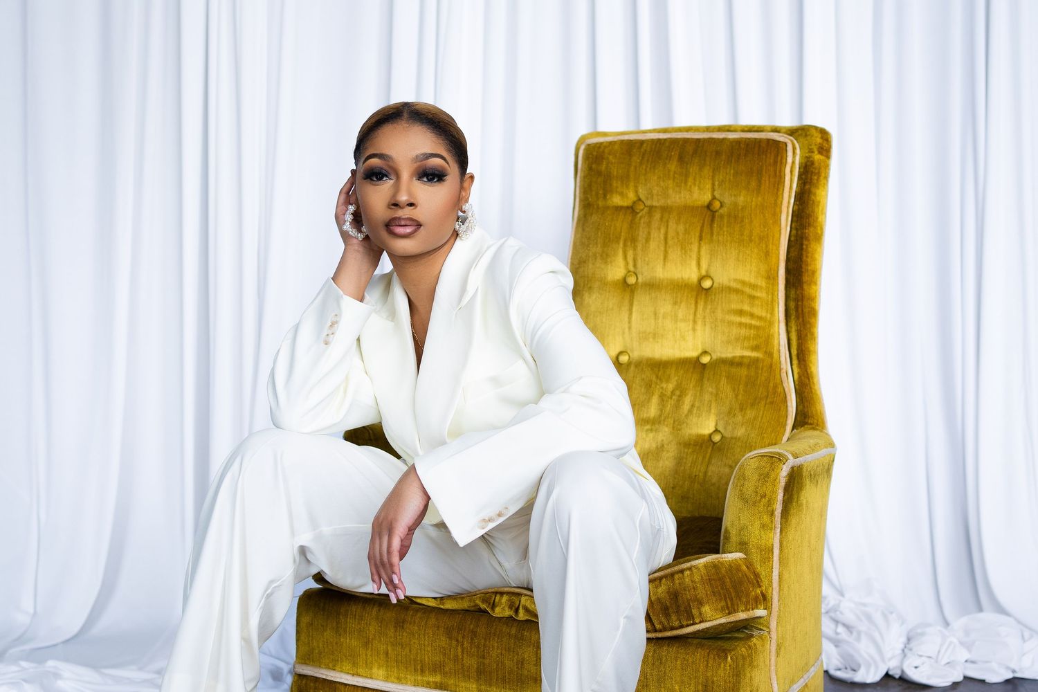 Fashion portrait in white suit seated on luxurious gold velvet chair against white backdrop.