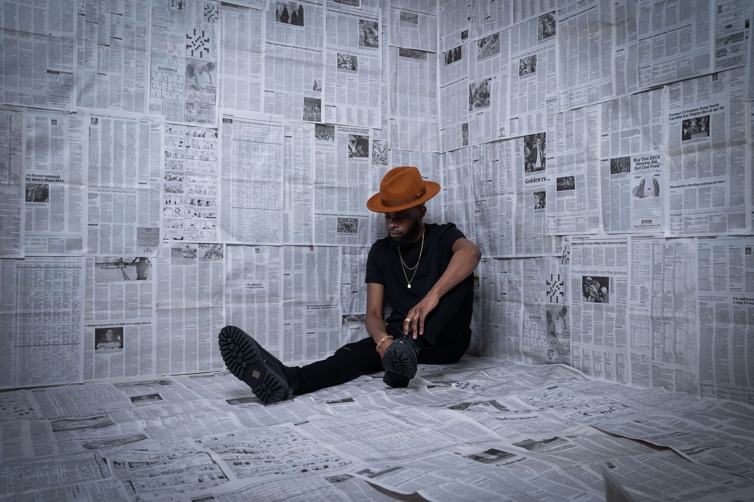 Figure in casual attire sits on floor against newspaper-covered walls.