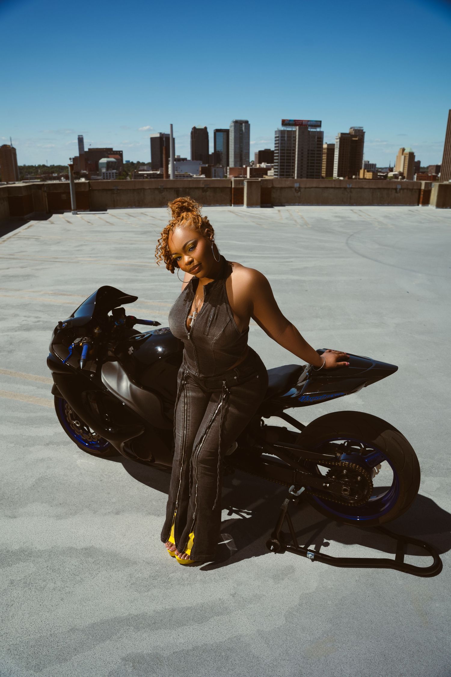 A woman in a dark jumpsuit poses beside a black motorcycle on a rooftop with a city skyline backdrop.