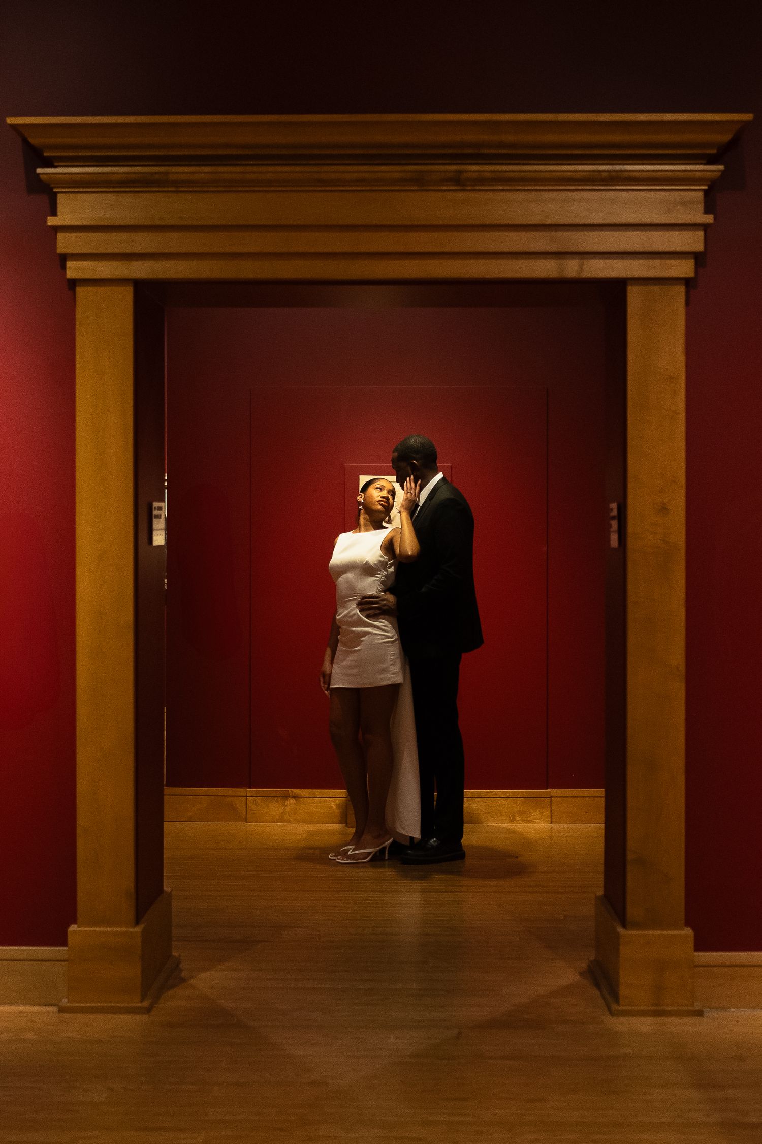 Dramatic lighting captures couple's tender moment through wooden archway in museum.